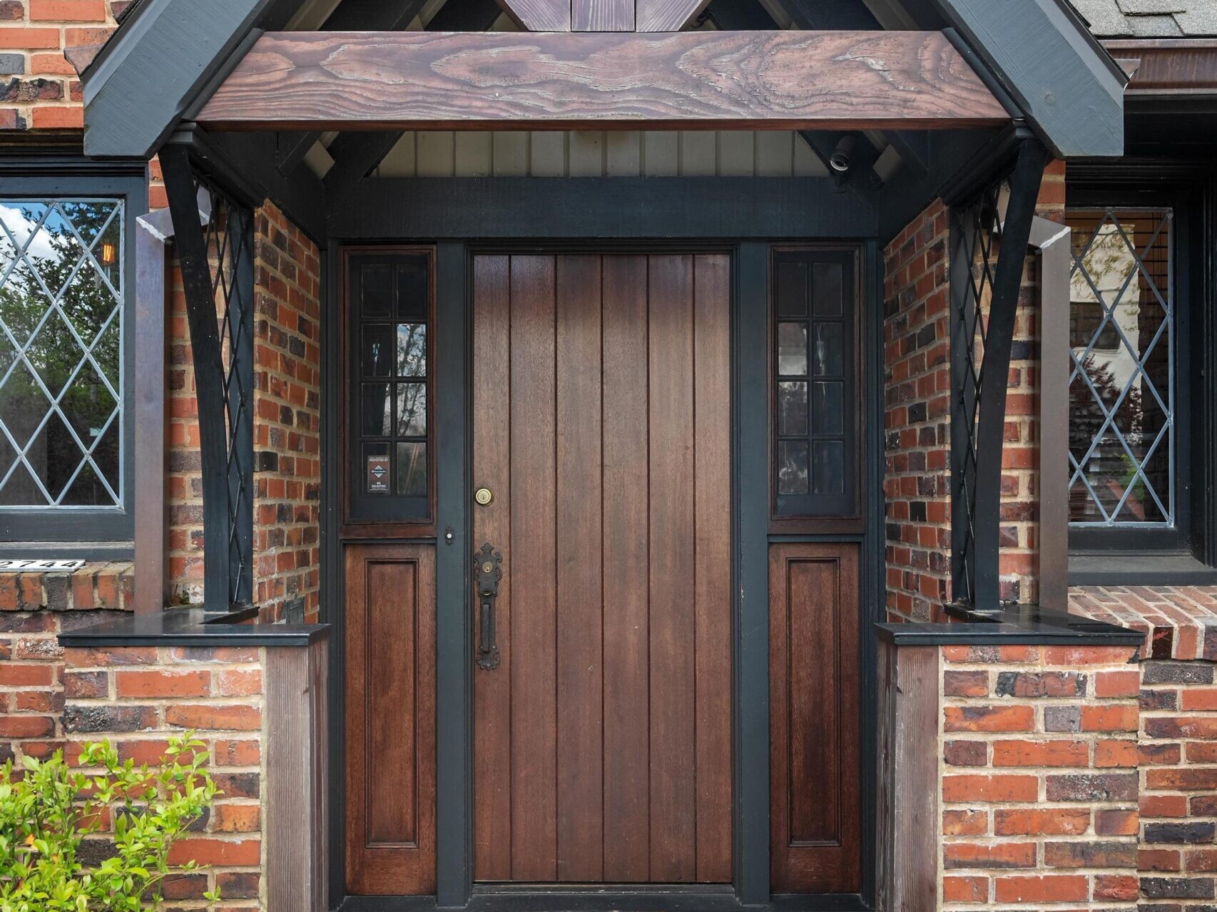 A wooden front door with side panels and a “Welcome” doormat sits under a covered entryway with dark wood beams on a brick house. Diamond-patterned windows and small green bushes frame the entrance.