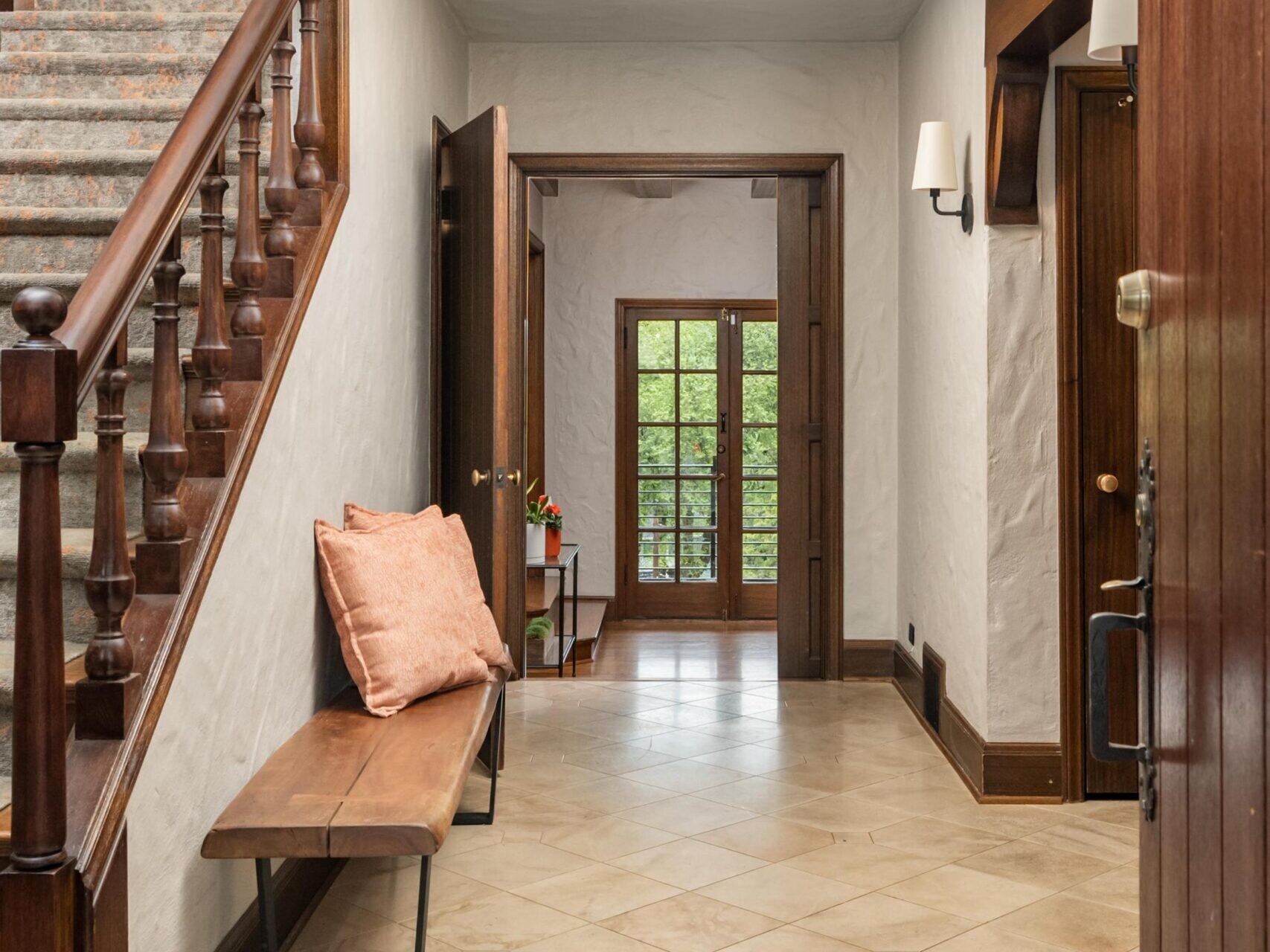 A hallway with tiled floors, a wooden staircase on the left, a bench with peach pillows, and double French doors at the end opening to a view of greenery outside.