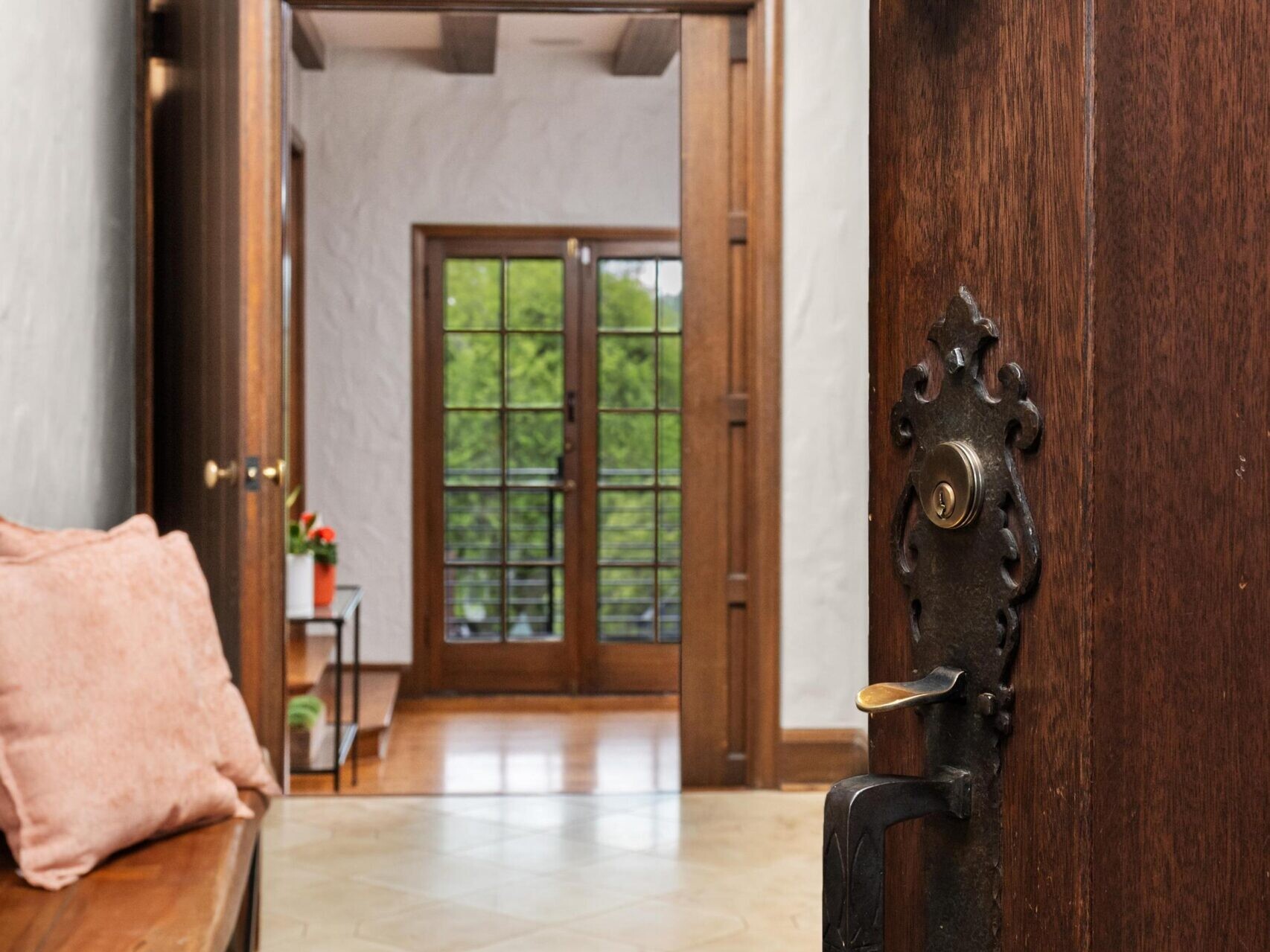 A partially open wooden door with an ornate handle reveals a hallway with tile flooring, a wooden bench with a pink cushion, and large windows overlooking greenery.