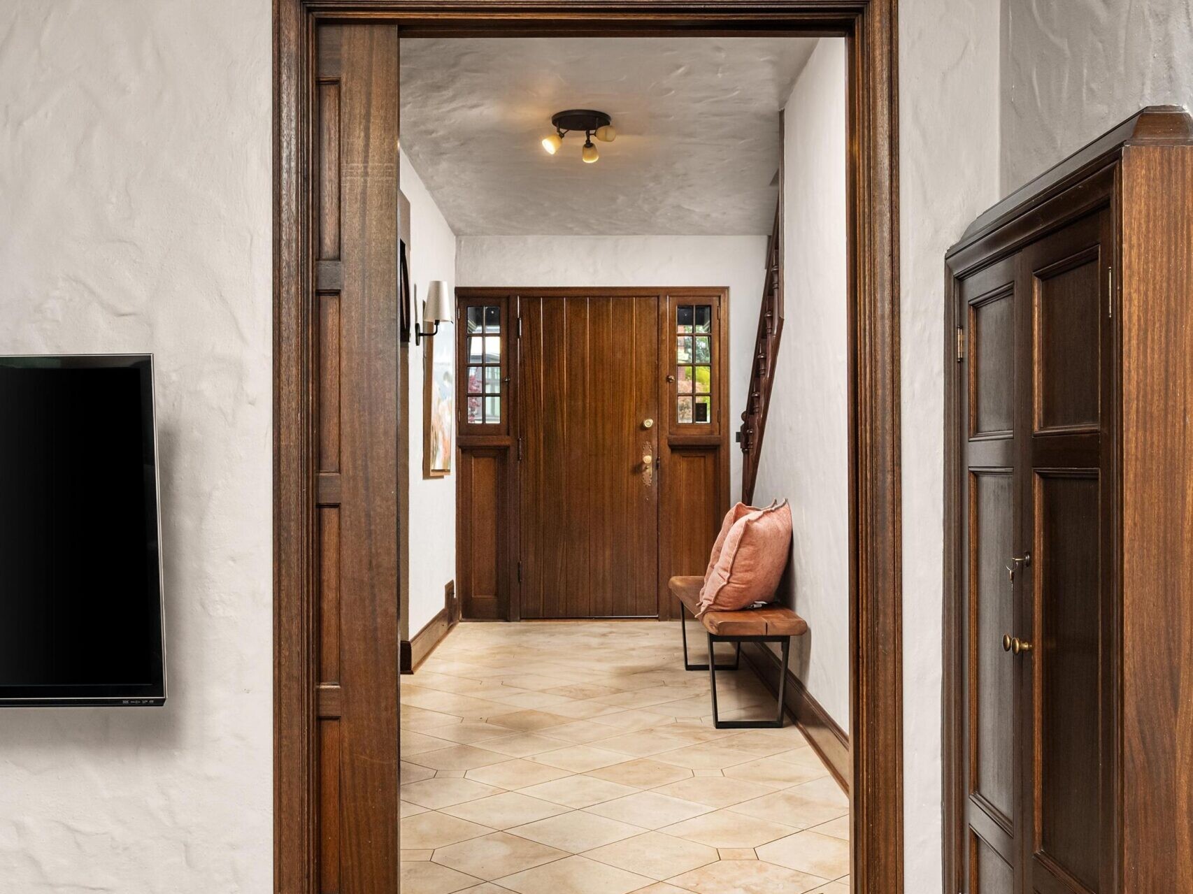A view from a living room into a hallway with wooden trim, white textured walls, and exposed ceiling beams. The hallway features a wooden door, tiled floor, a bench with a pillow, and a wall sconce.