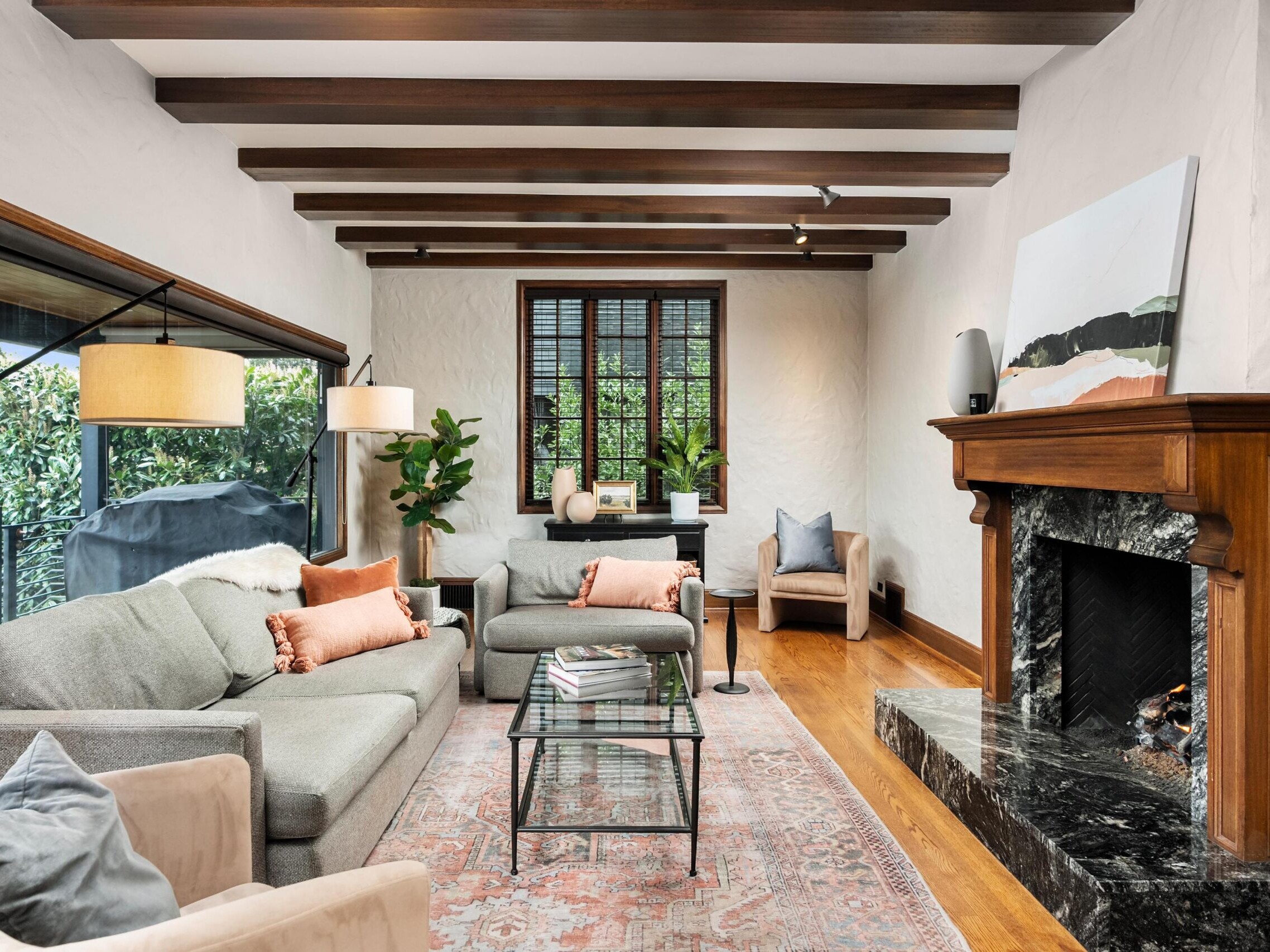 A cozy living room with exposed wooden ceiling beams, gray sofas, a marble fireplace, large window, indoor plants, glass coffee table, and neutral-toned decor with peach and beige accents.