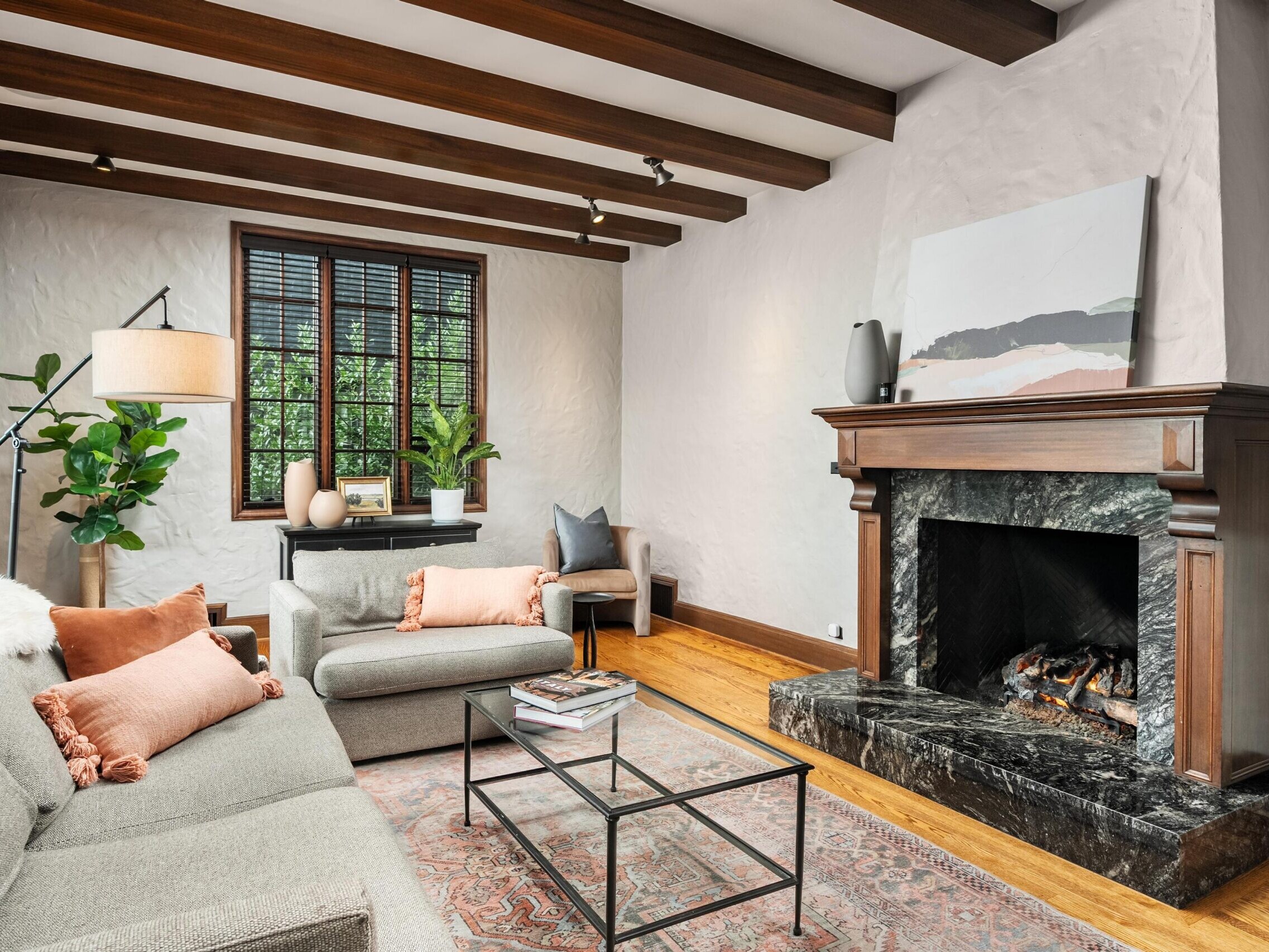 A cozy living room features a gray sofa, armchair, and a black marble fireplace. Wooden beams line the ceiling, and a large window lets in natural light. Decorative plants and pastel accents add warmth to the space.