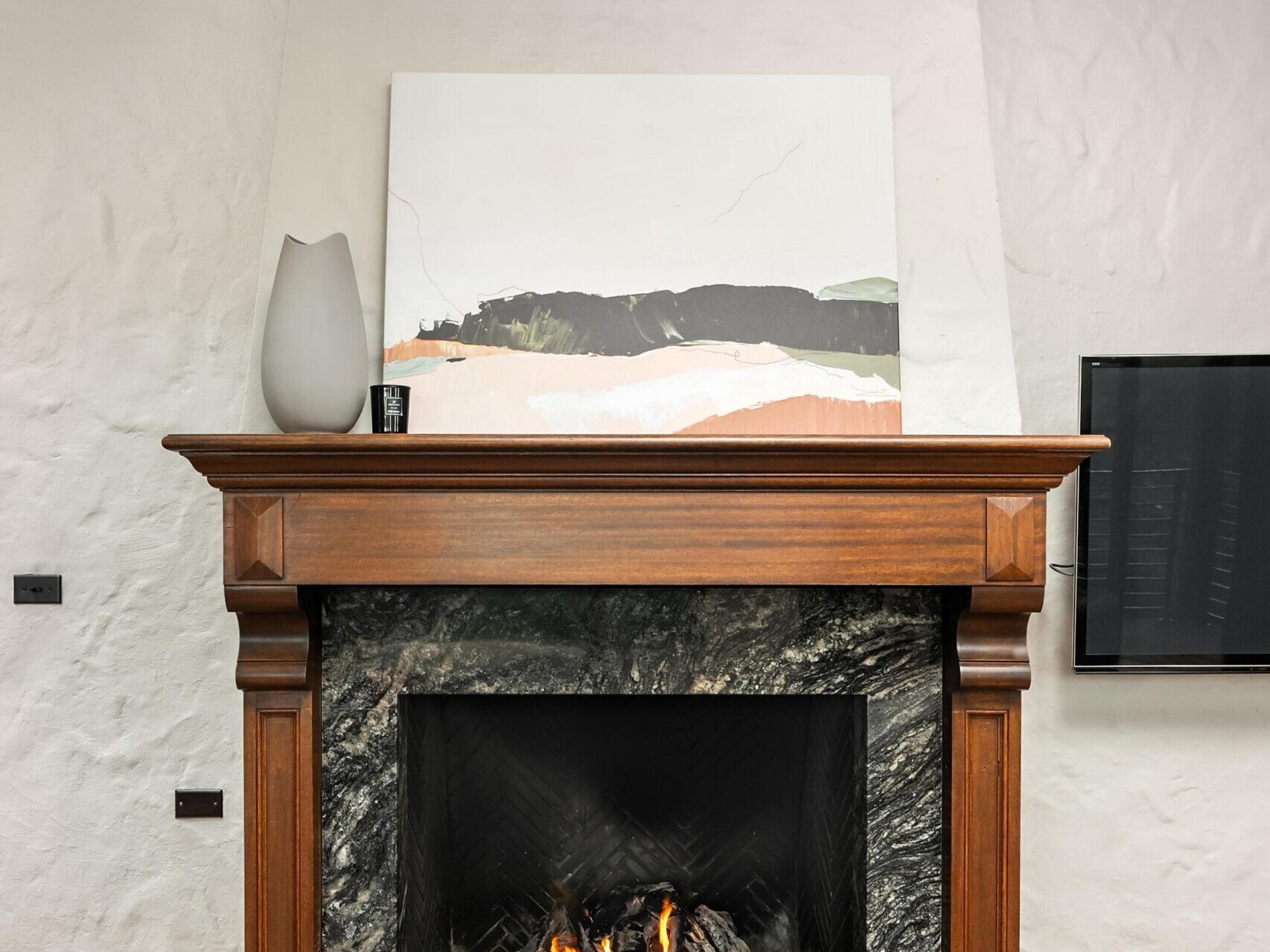 A modern fireplace with a dark marble surround and wooden mantel, lit with a small fire. A large abstract painting and a gray vase sit on the mantel. The room has white textured walls and exposed wooden ceiling beams.