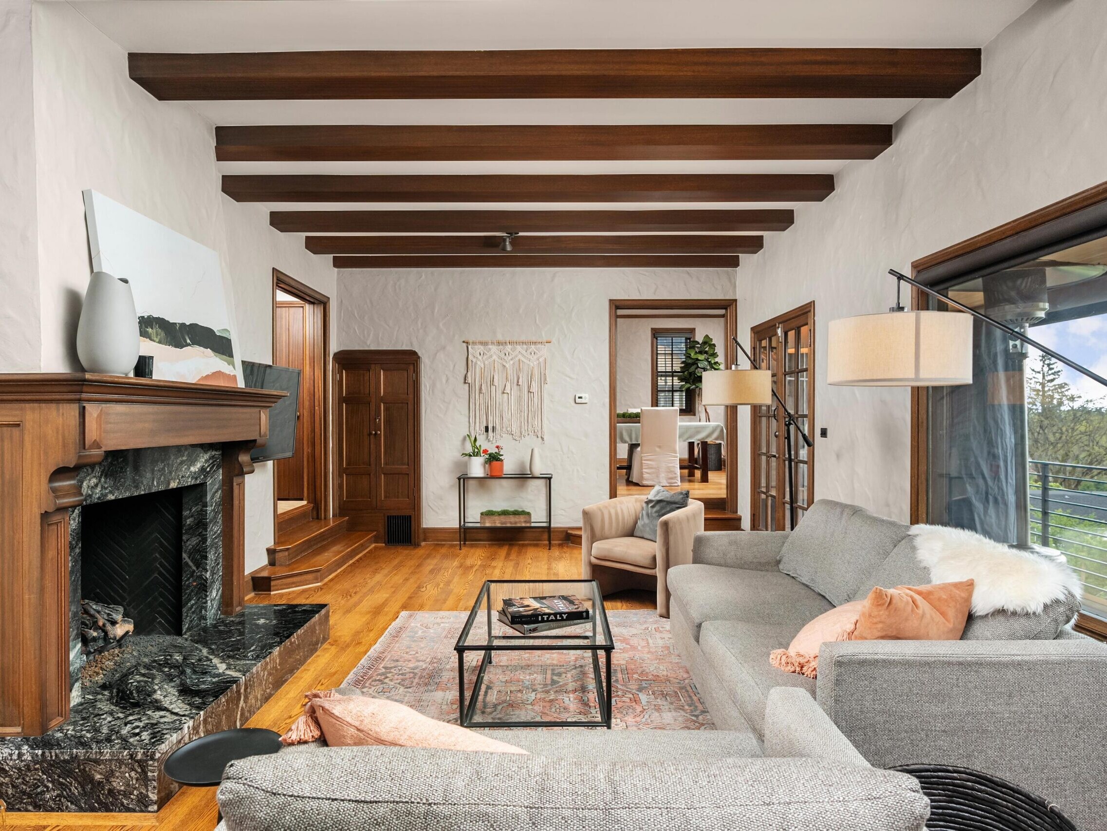 A cozy living room with wood ceiling beams, a gray sofa, armchairs, a marble fireplace, a rug, and a coffee table. Glass doors open to a balcony with a scenic outdoor view.