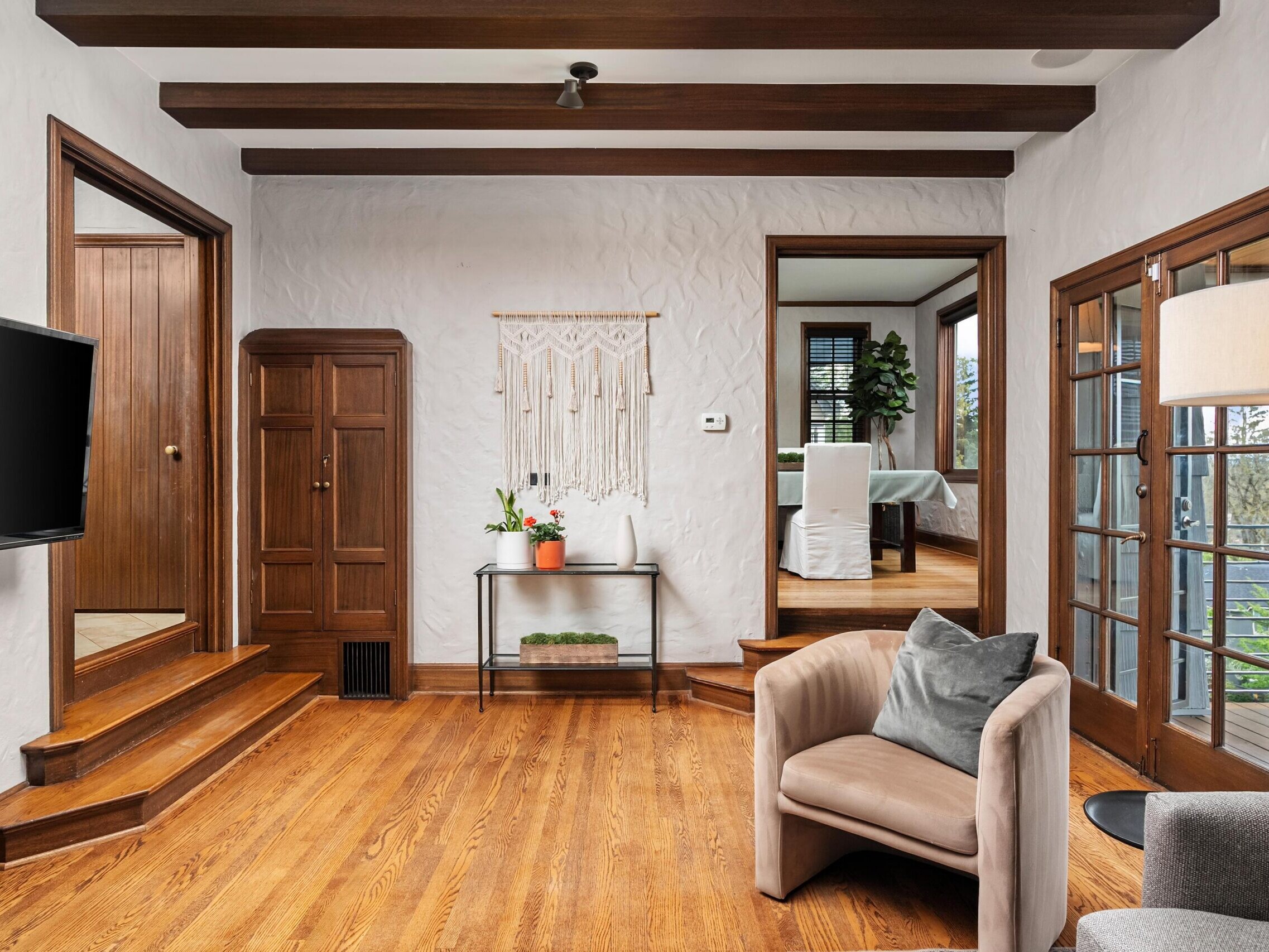A cozy living room with wooden floors, ceiling beams, and white textured walls. There are wooden doors, a beige armchair with a gray pillow, a wall-mounted TV, and a small table with plants and a macramé wall hanging.