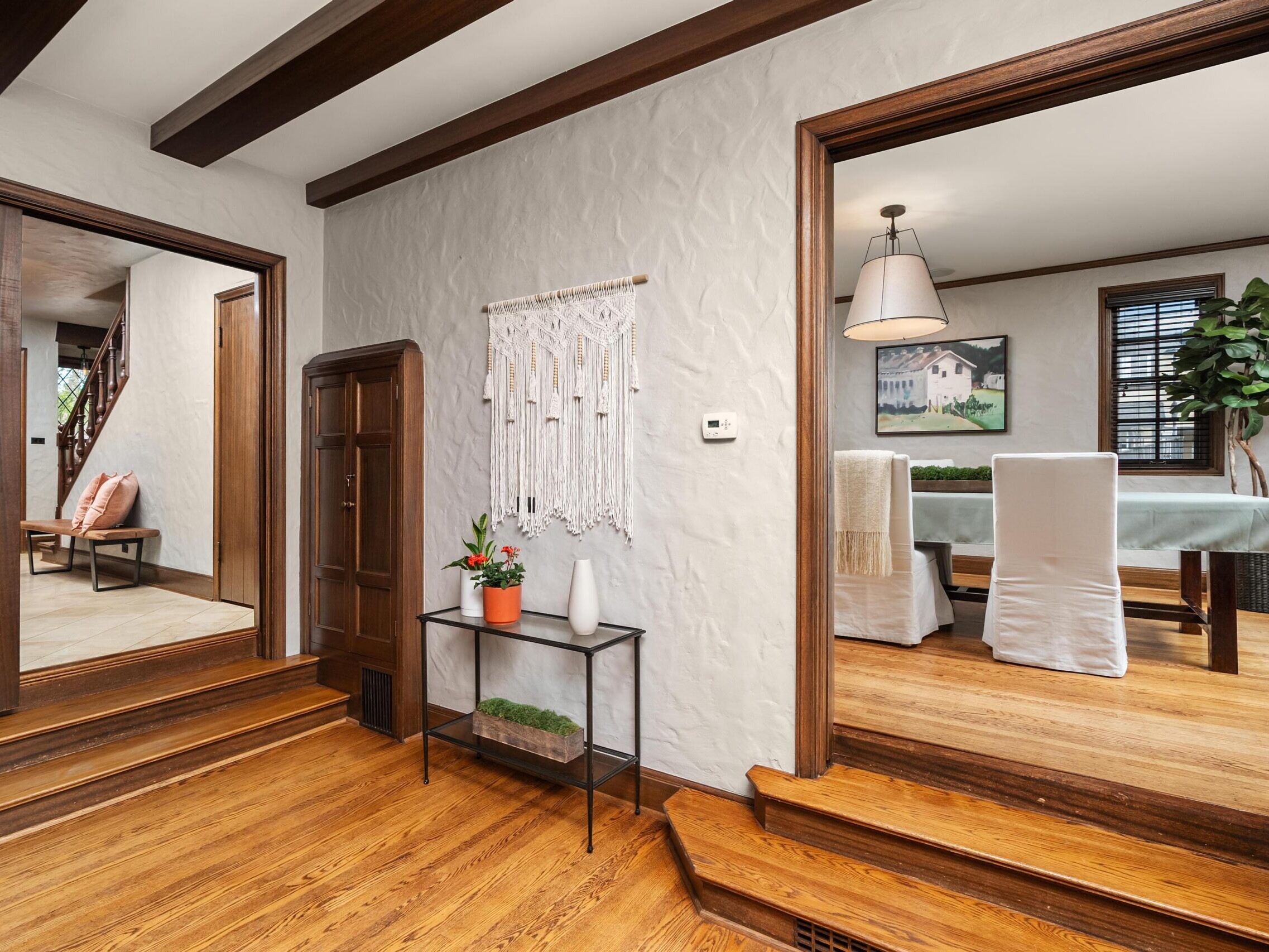 A hallway with wooden floors and exposed ceiling beams leads to a living area with chairs on the left and a dining room with a table and white chairs on the right. A console table with decor sits against a textured wall.