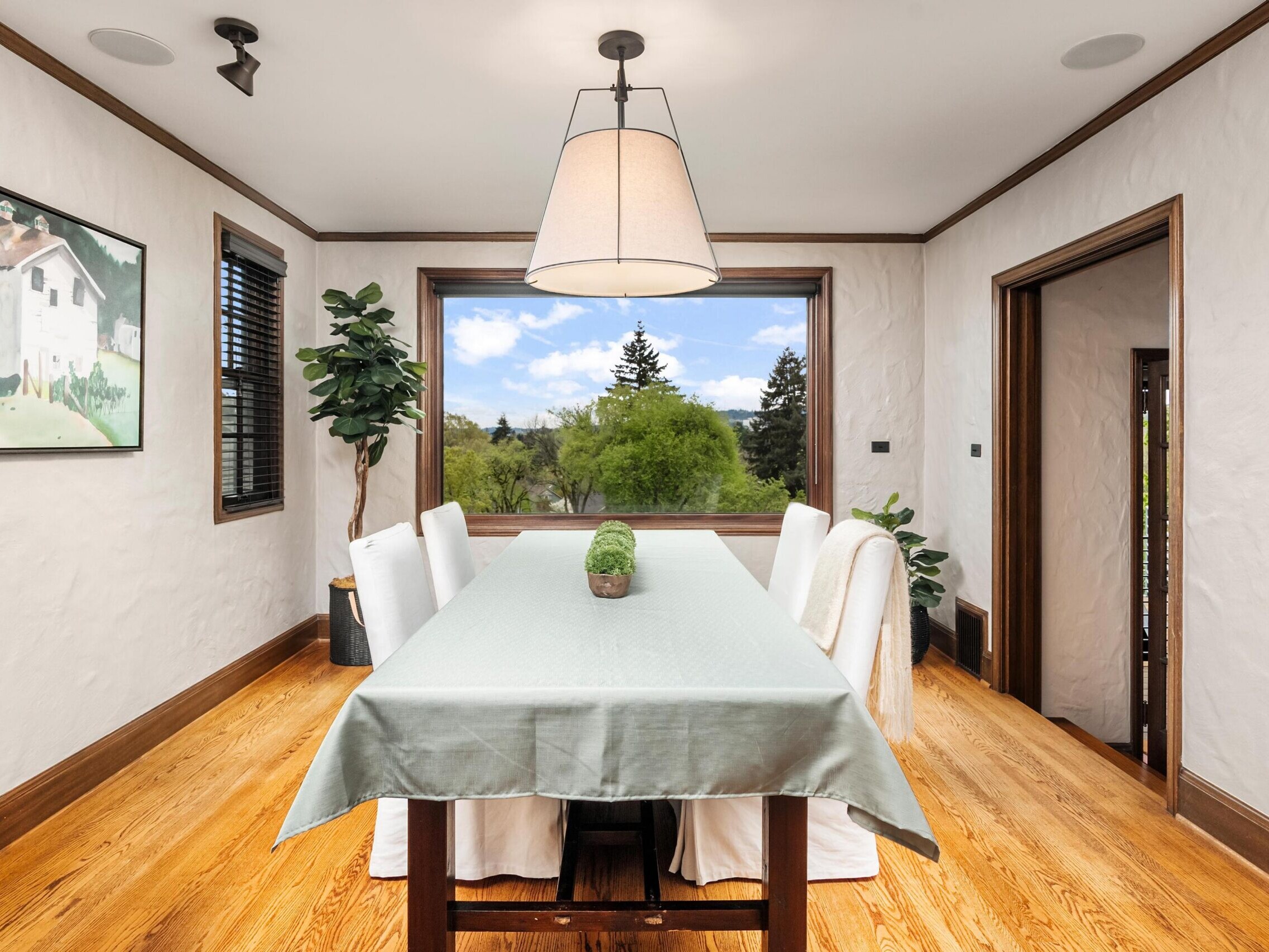 Bright dining room with a large window showcasing a scenic outdoor view, a long wooden table covered with a light tablecloth, white chairs, a plant centerpiece, wall art, and wooden floors.