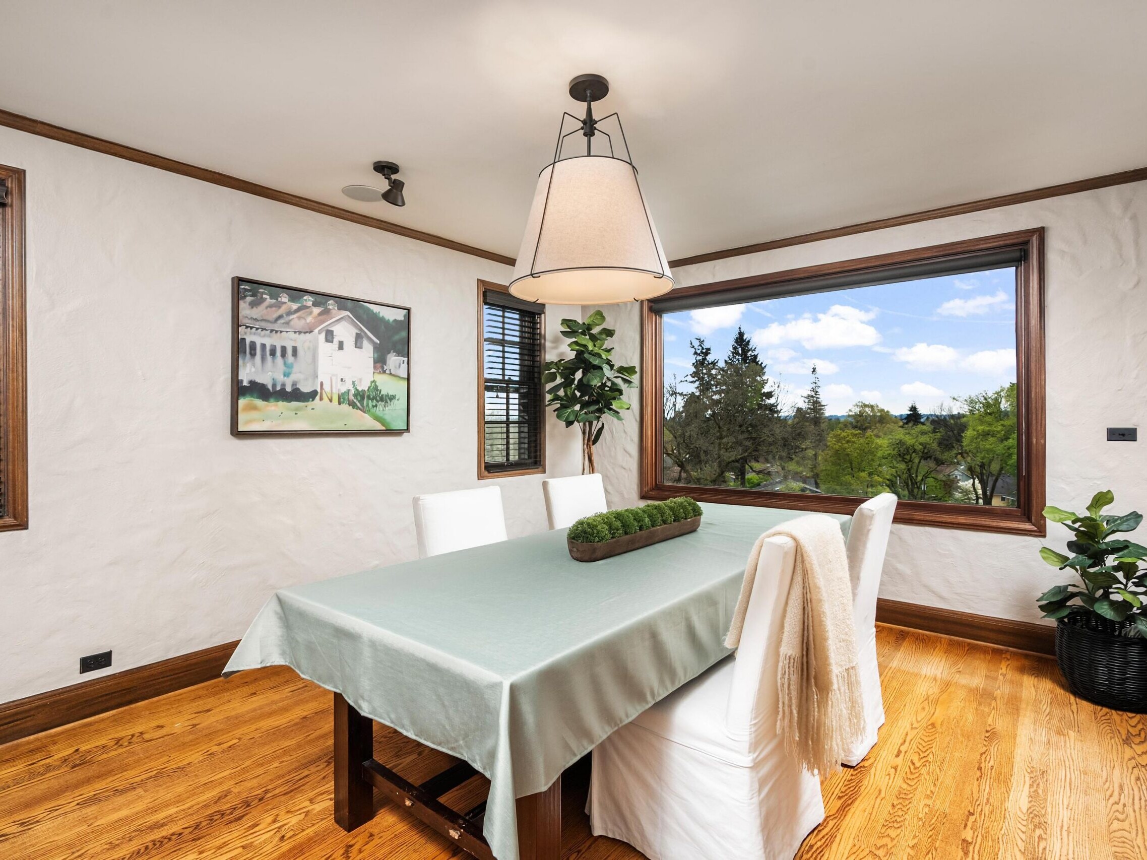 A bright dining room with a wooden floor, a table covered by a pale green cloth, four white chairs, a large window overlooking trees, a potted plant, a landscape painting, and a modern pendant light overhead.