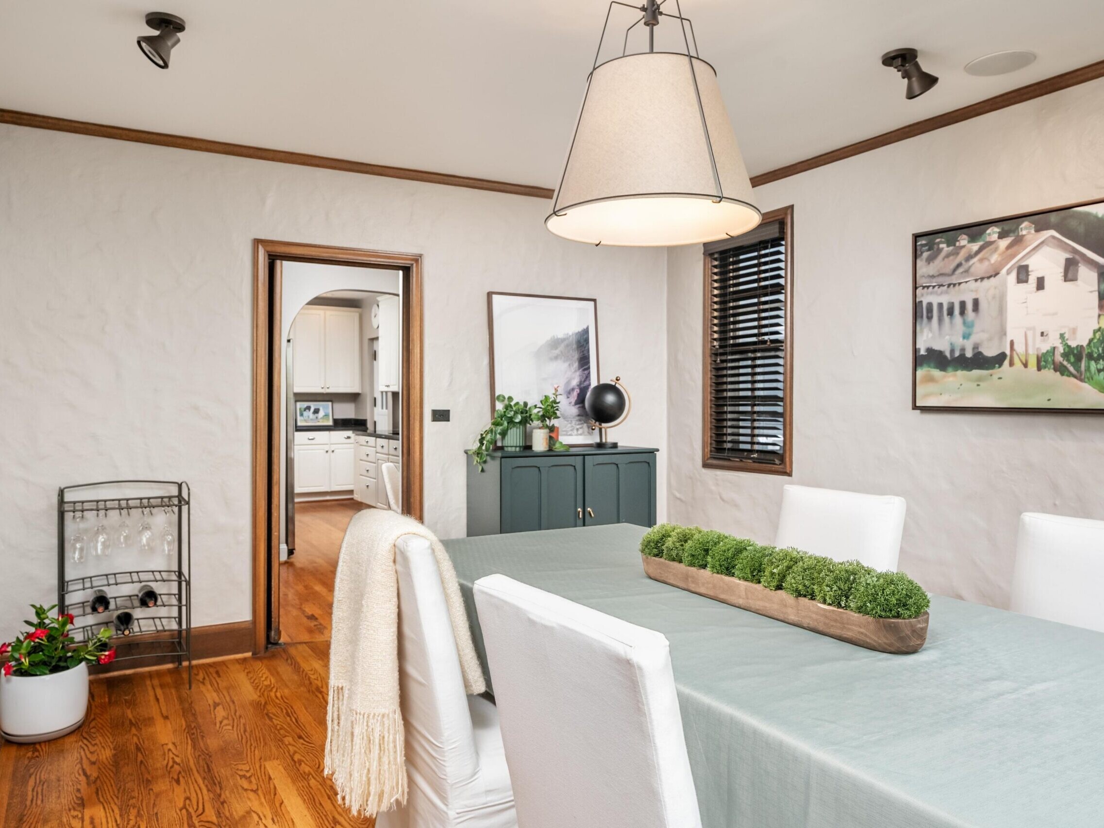 A dining room with a table covered by a light blue cloth, white upholstered chairs, a rectangular planter centerpiece, modern pendant light, wall art, wooden floors, and a view into a bright kitchen.