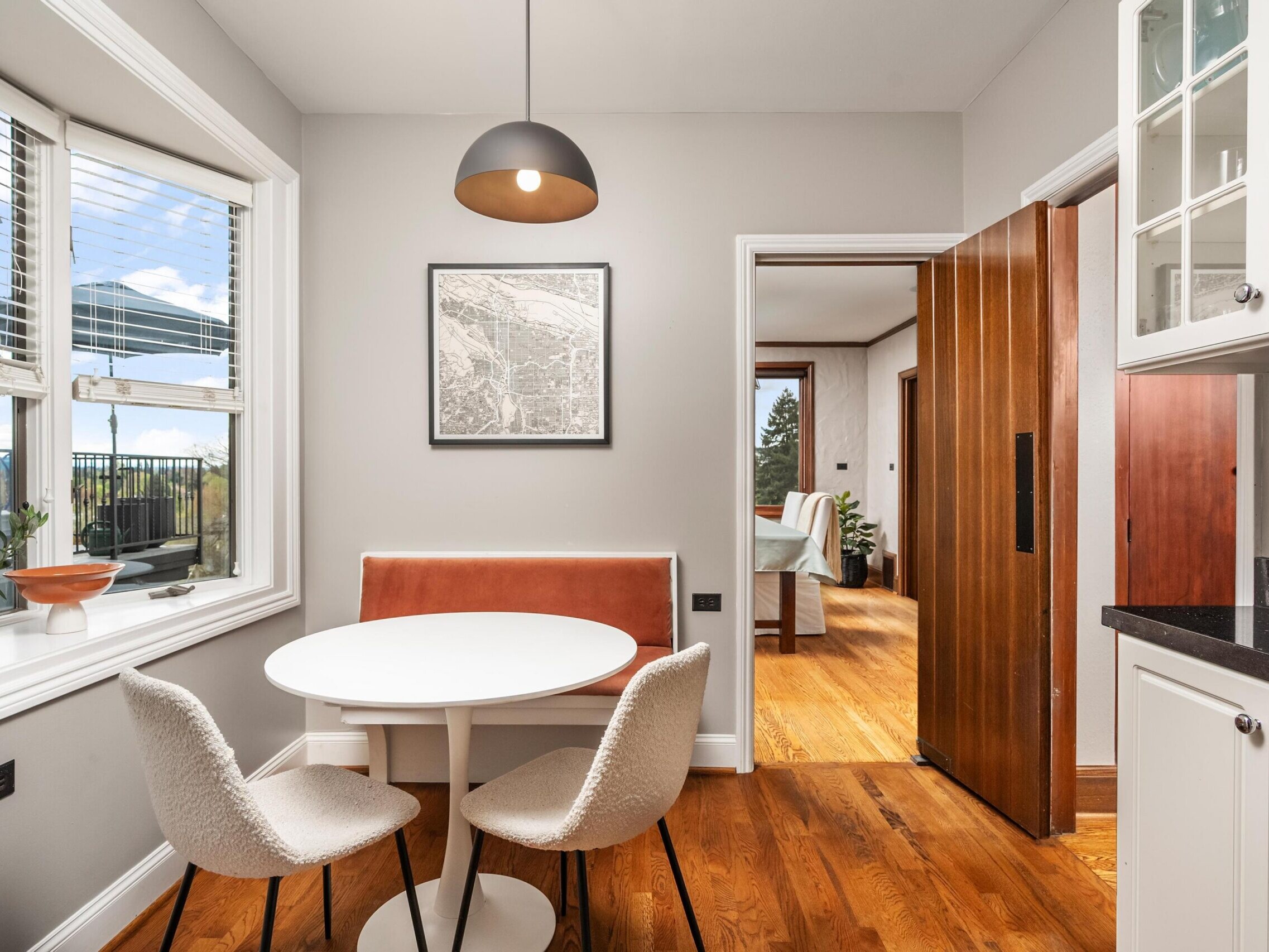 A small dining nook with a round white table, two upholstered chairs, a built-in bench, and a hanging lamp sits by a window in a modern kitchen with wood floors and sliding doors to a sunlit living area.