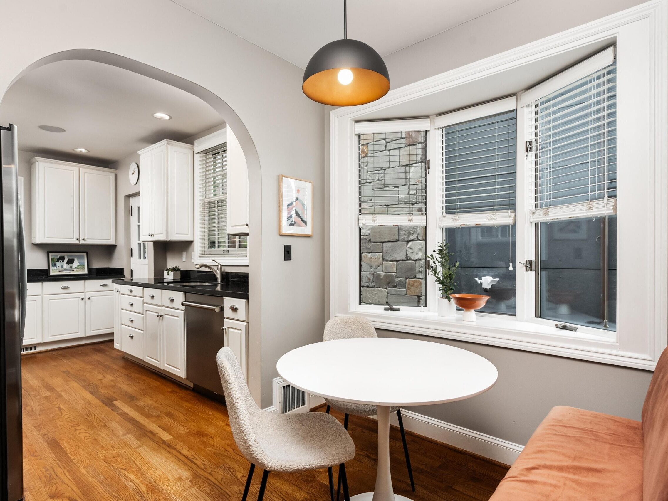 A modern kitchen and dining nook with wood floors, white cabinets, stainless steel appliances, and a round table with two chairs by a large bay window. A pendant light hangs above the table.