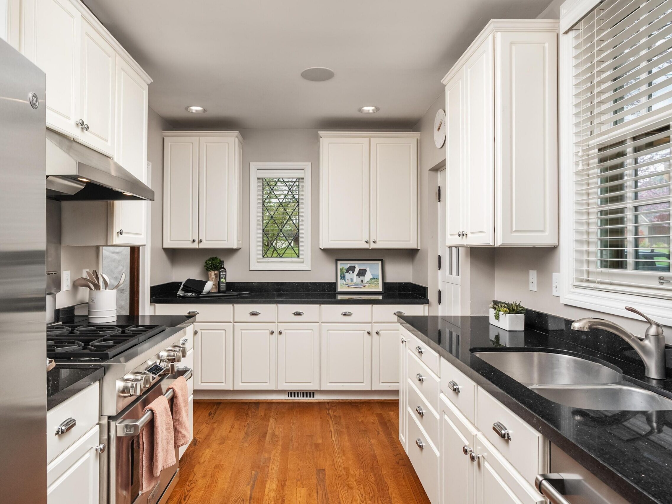 Modern kitchen with white cabinets, black countertops, stainless steel appliances, a double sink, hardwood floors, a small window above the counter, and a window with blinds on the right. Decor items and a plant are on the counters.
