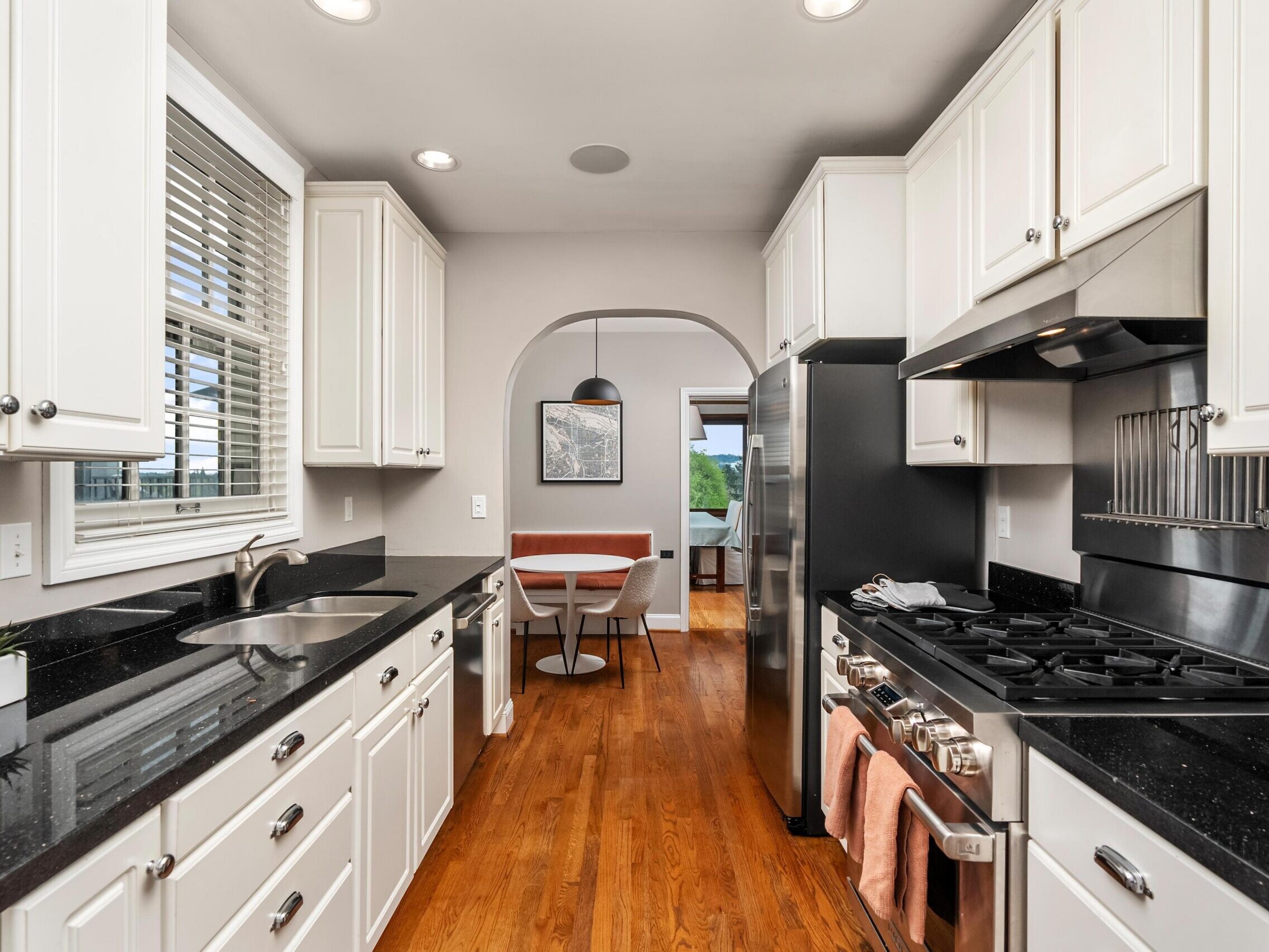 Modern galley kitchen with white cabinets, black countertops, stainless steel appliances, and wood floors. An arched doorway leads to a dining nook with a round table and a window with greenery visible outside.