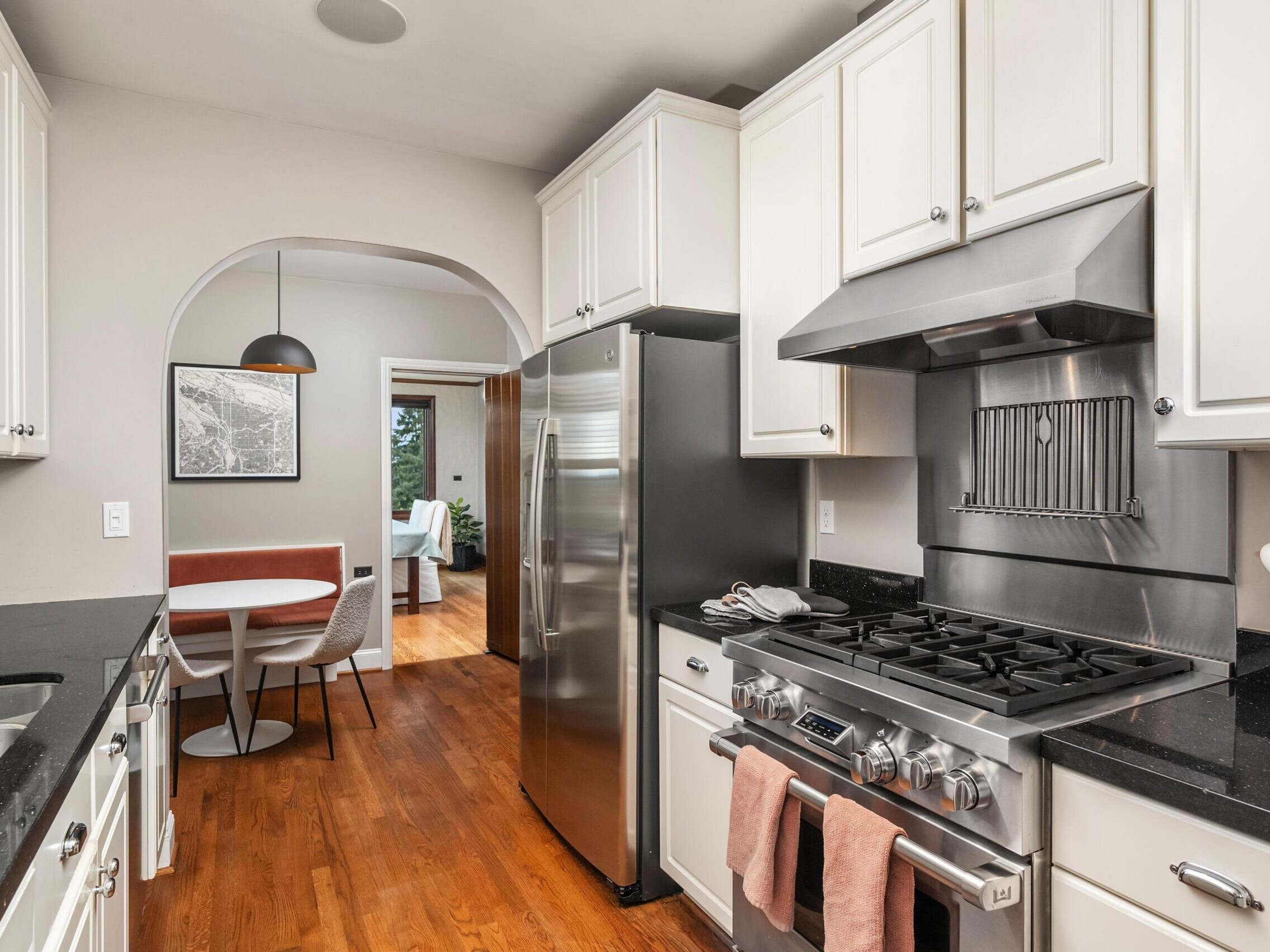 Modern kitchen with white cabinets, stainless steel appliances, black countertops, and wooden floor. An archway leads to a dining area with a round table and chairs, and a pendant light above.