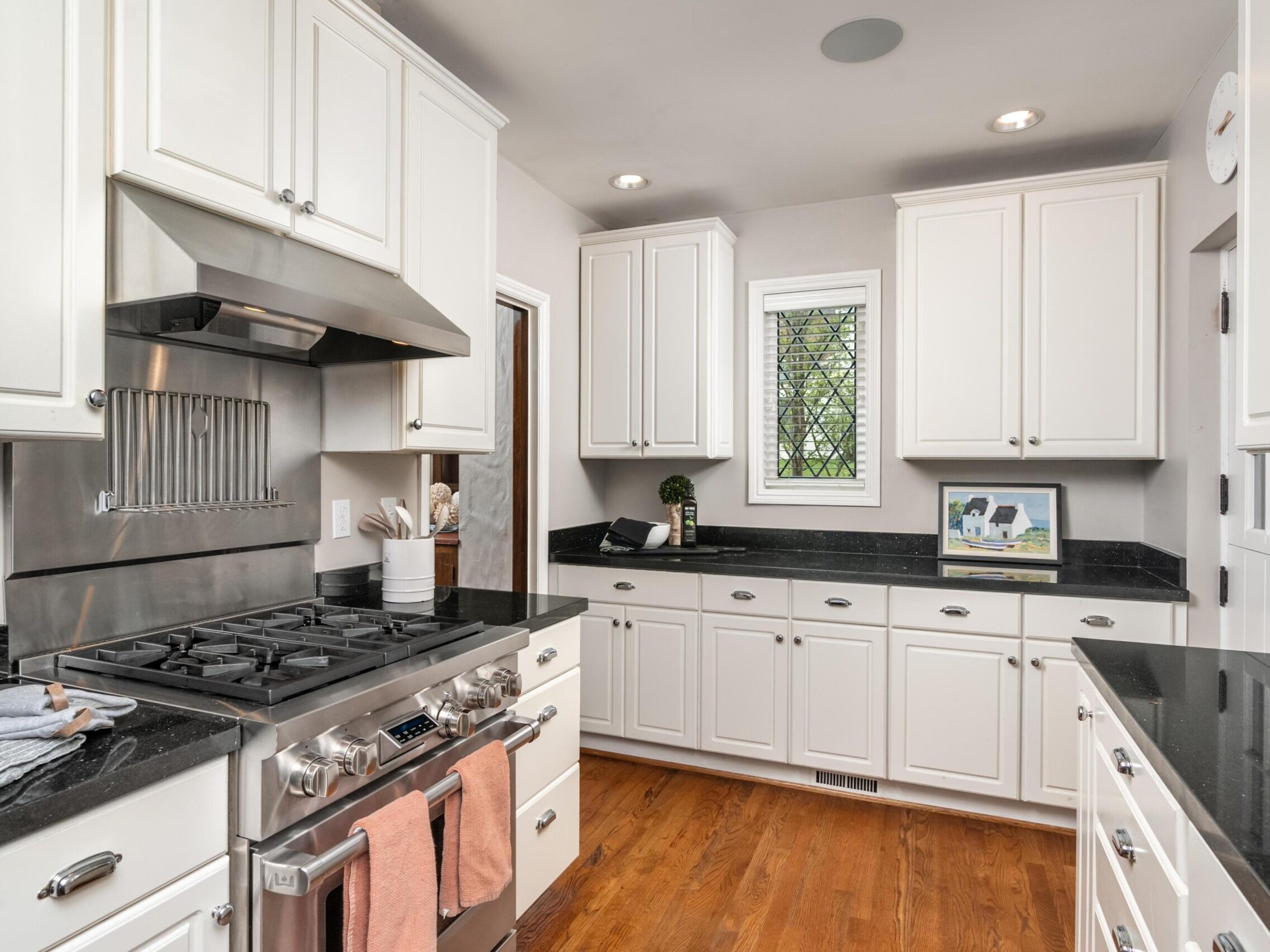 A modern kitchen with white cabinets, black countertops, stainless steel appliances, a gas stove with pink towels, and wooden flooring. A small window with a lattice design lets in natural light.