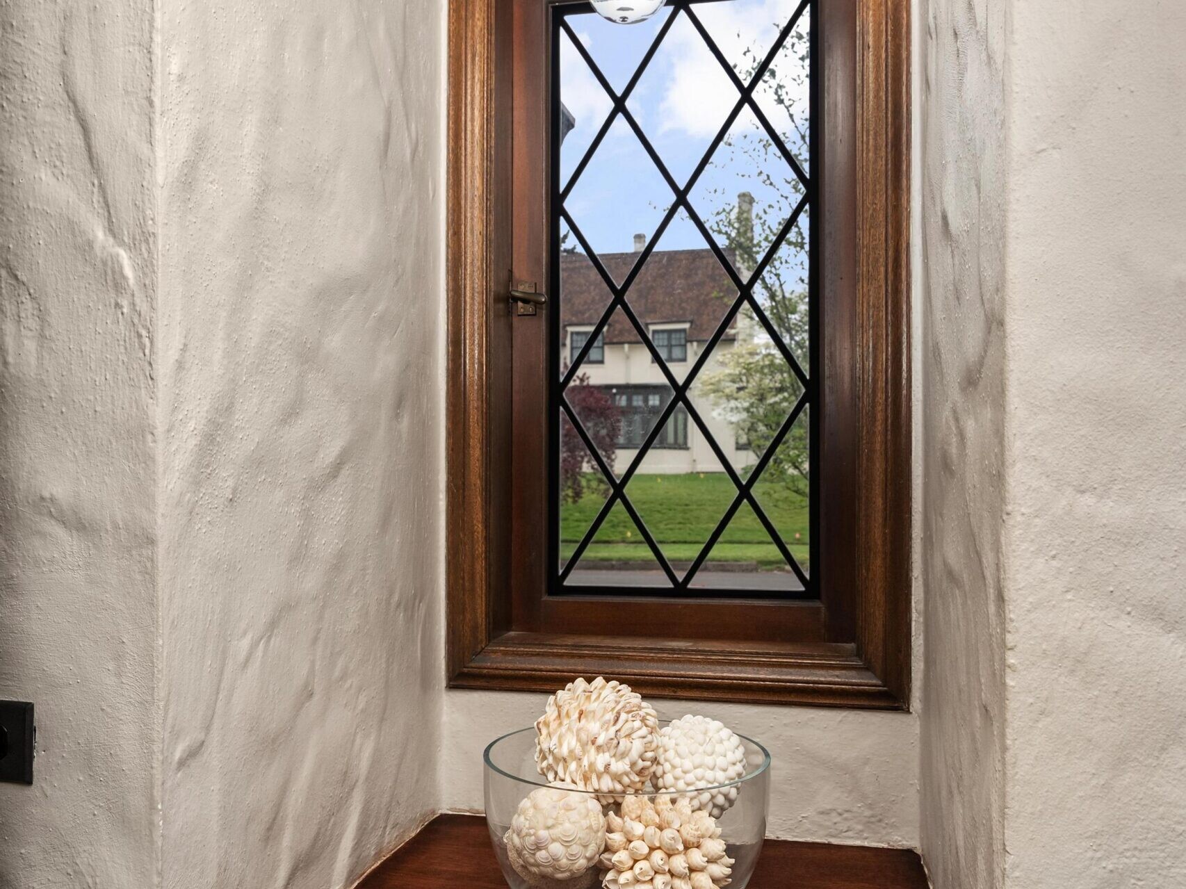 A small window with a wooden frame and diamond-patterned glass overlooks a house and greenery. Beneath the window, a wooden surface holds a glass bowl filled with decorative white spheres. A single light fixture hangs above.
