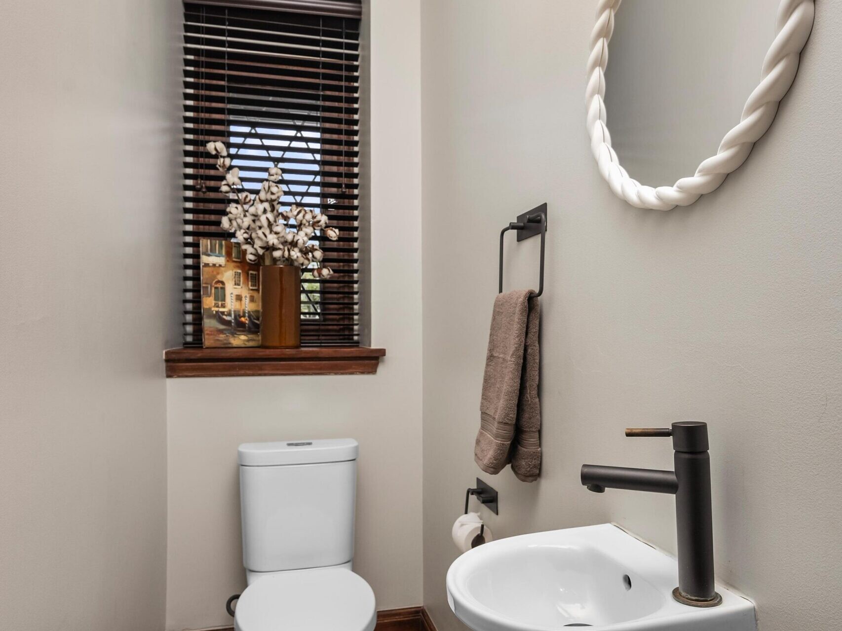 A small, modern bathroom with a white toilet, a round mirror above a small white sink, a black faucet, towel holder with two towels, a window with wooden blinds, and a vase of flowers on the window sill.