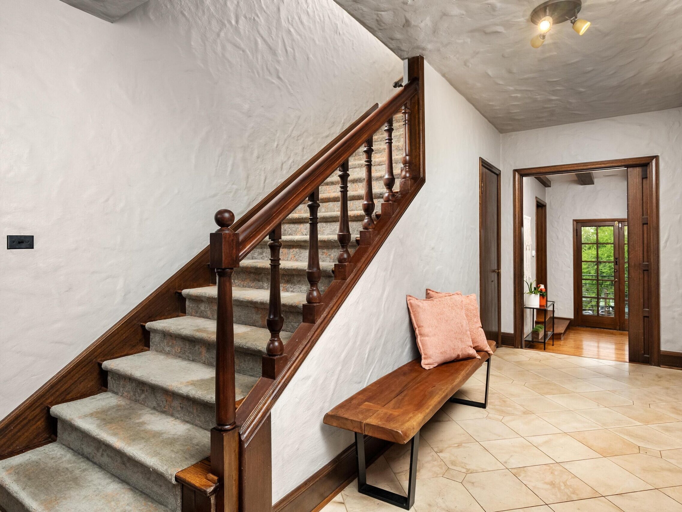 A hallway with tiled floors, a wooden staircase with carpeted steps, a wooden bench with a pink pillow, and doorways leading to other rooms with natural light and greenery visible outside.
