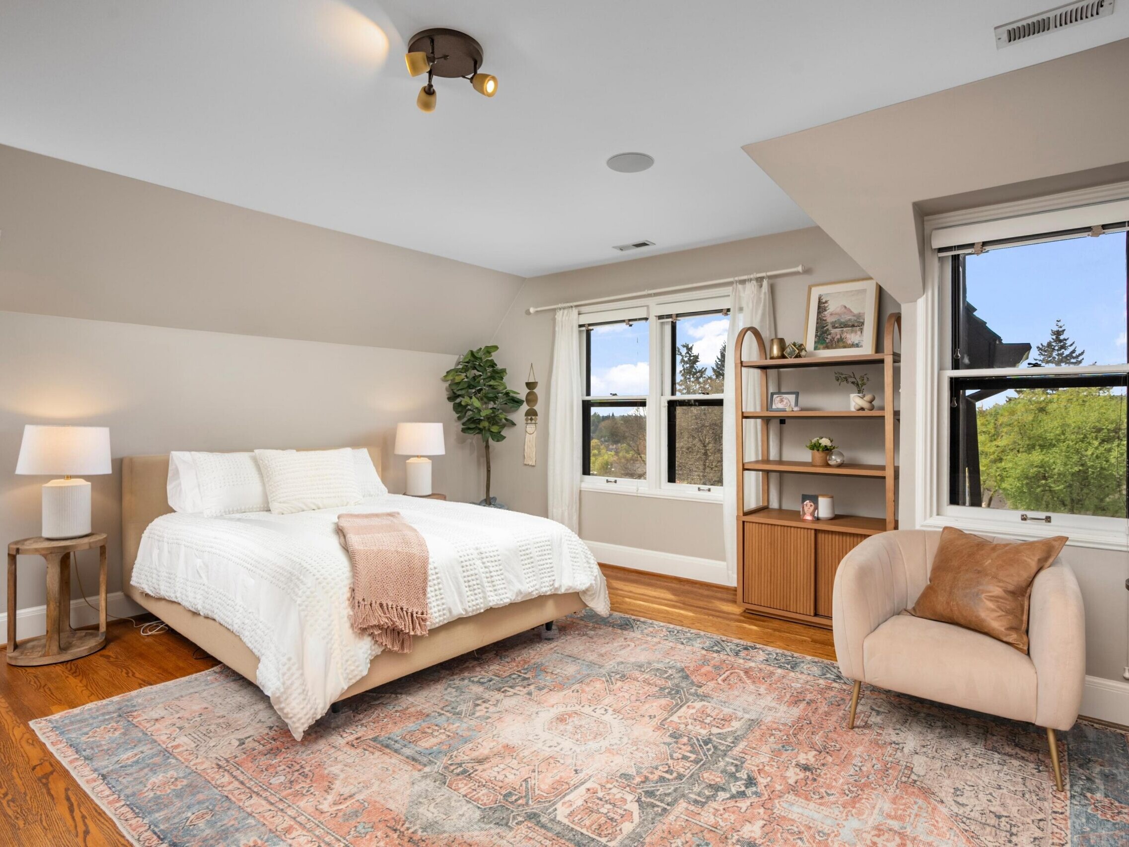 A cozy bedroom with a bed, white bedding, two bedside tables with lamps, a beige armchair, a wooden bookshelf, a large patterned rug, a potted plant, and two windows showing trees outside.