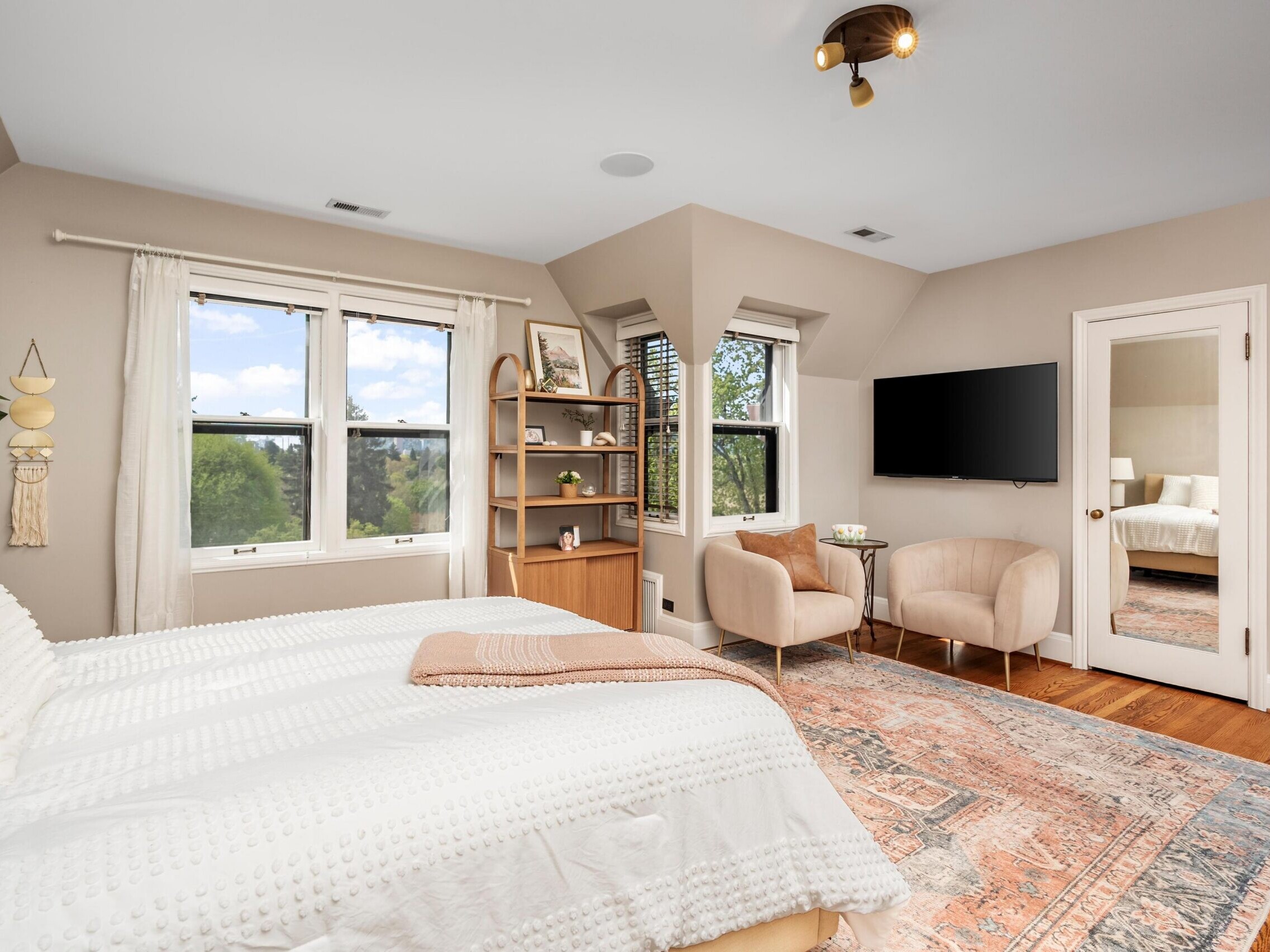 Bright, cozy bedroom with a large bed, two beige chairs, a wall-mounted TV, bookshelves, and large windows letting in natural light. Decor includes woven wall hangings, a potted plant, and a patterned rug.