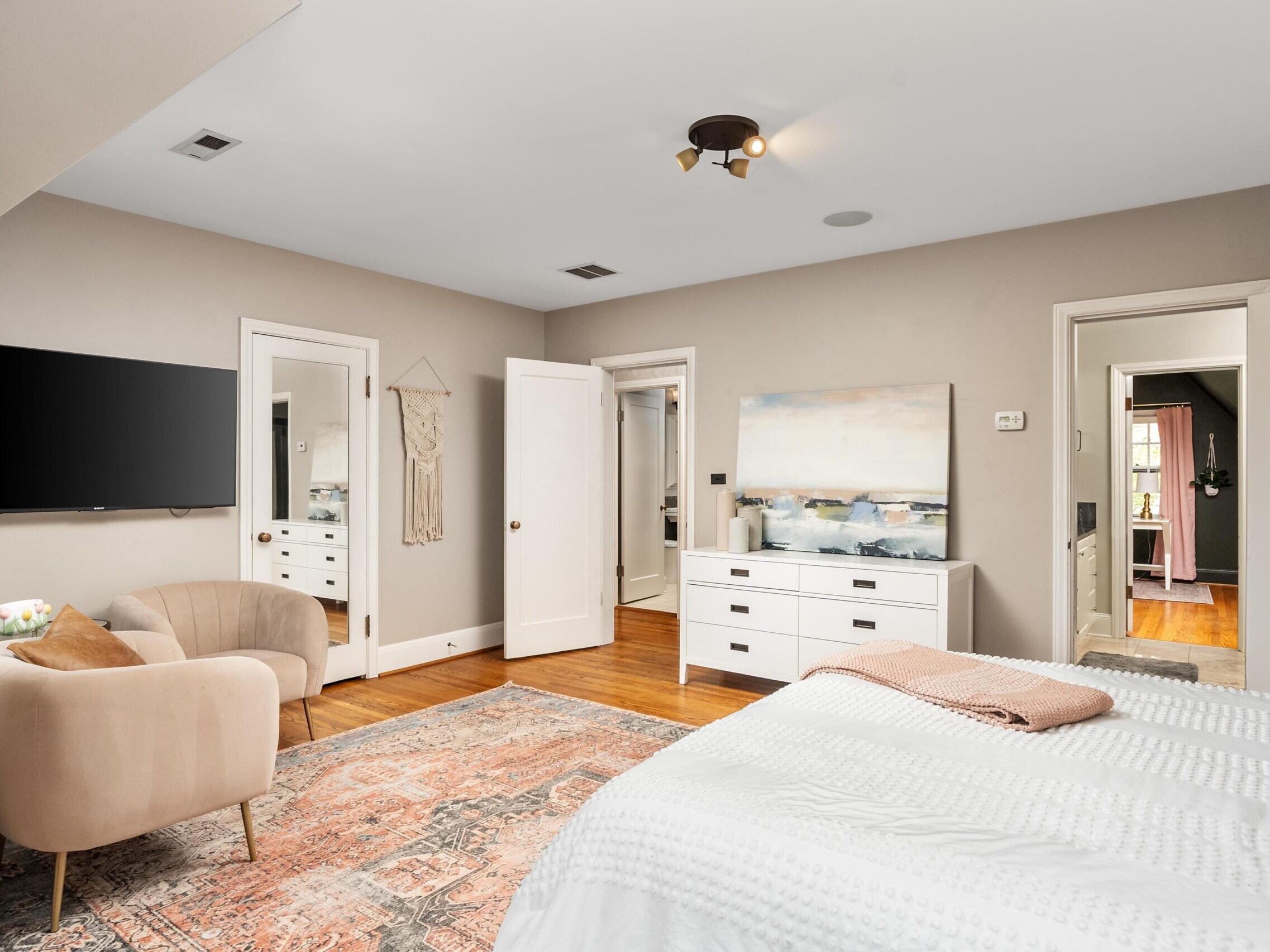 Bright bedroom with a large rug, white bed, beige armchair, wall-mounted TV, dresser with art above, double mirrors, and open doors showing adjoining rooms. The walls are light, and the decor is modern and cozy.