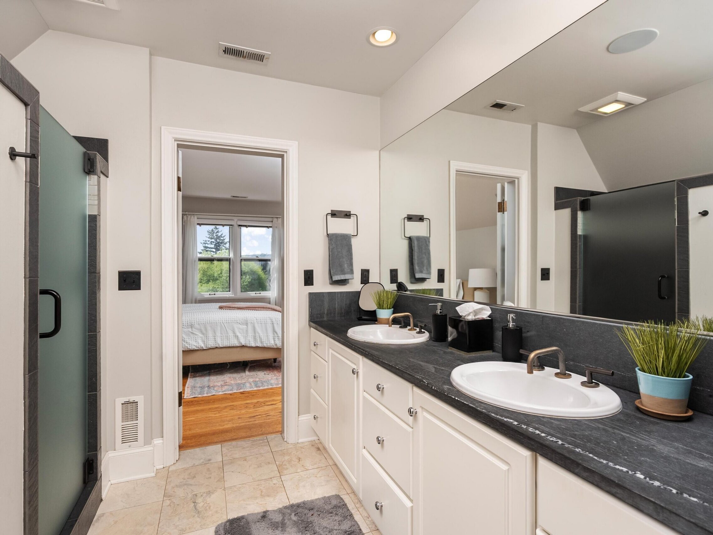 Modern bathroom with double sinks, large mirror, black countertop, and white cabinets. A glass shower is on the left, and an open door leads to a bright bedroom with large windows.