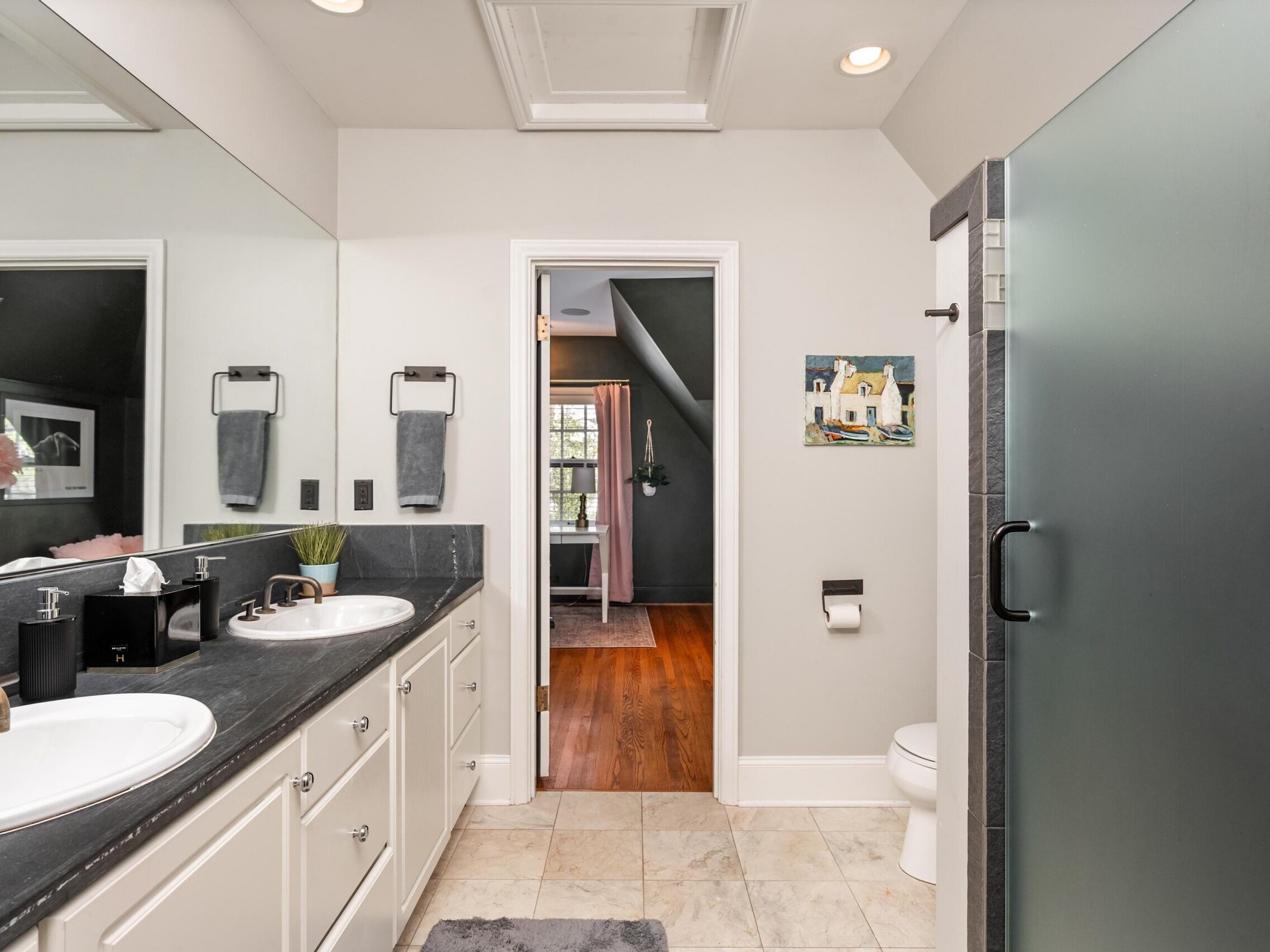 Modern bathroom with double sinks, black countertops, towels, frosted glass shower, toilet, and wall art. Door opens to a room with hardwood floor and large window with curtains letting in sunlight.