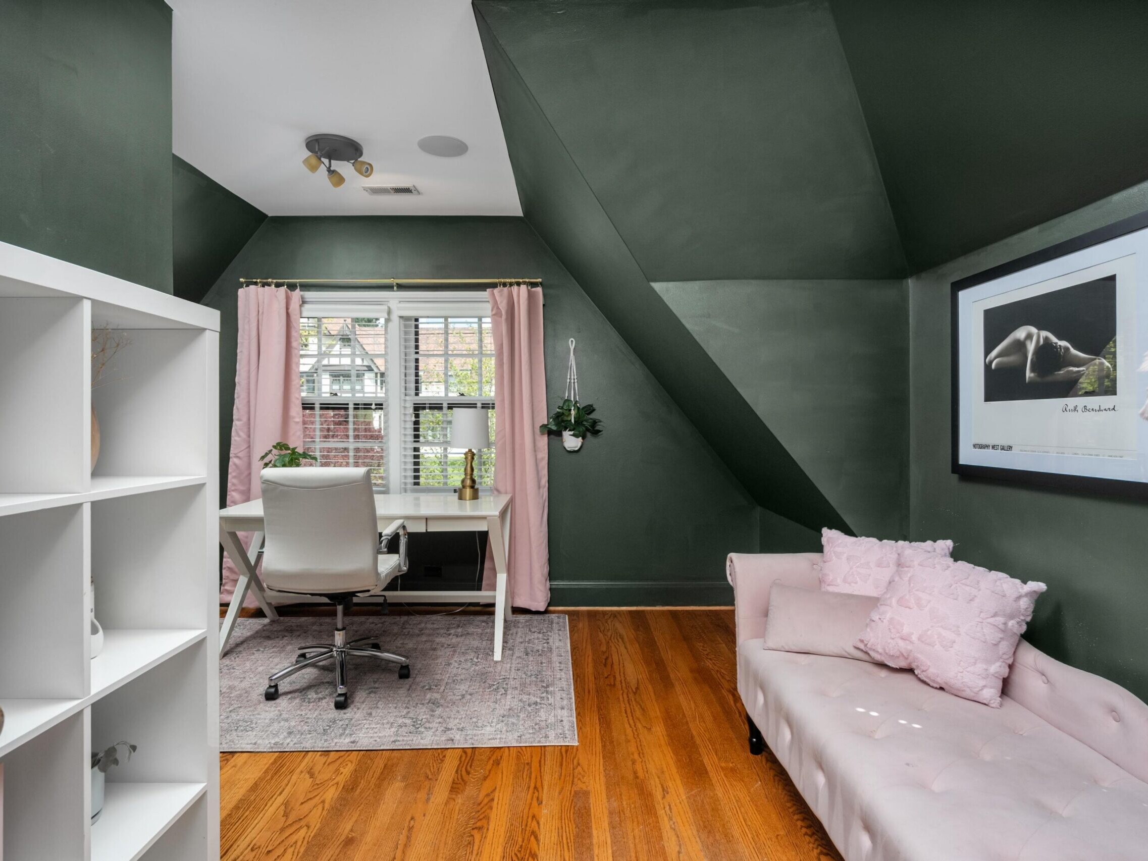 A home office with dark green walls, wooden floor, a white desk and chair by a window with pink curtains, a light pink sofa with pillows, and a bookshelf on the left. A framed photo hangs on the wall above the sofa.