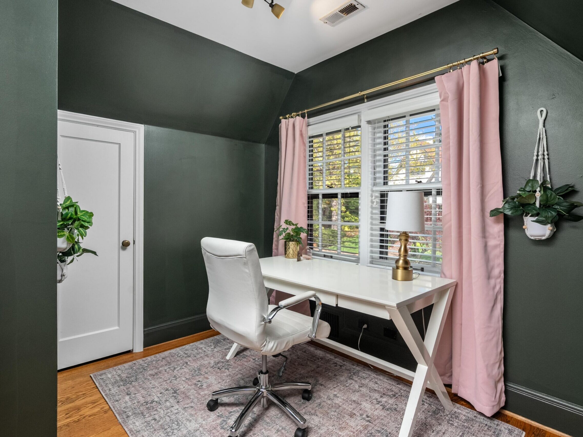 A home office with dark green walls, a white desk and chair, a gold lamp, and a window with white blinds and pink curtains. Hanging plants are on both sides of the window and a light purple rug covers the floor.