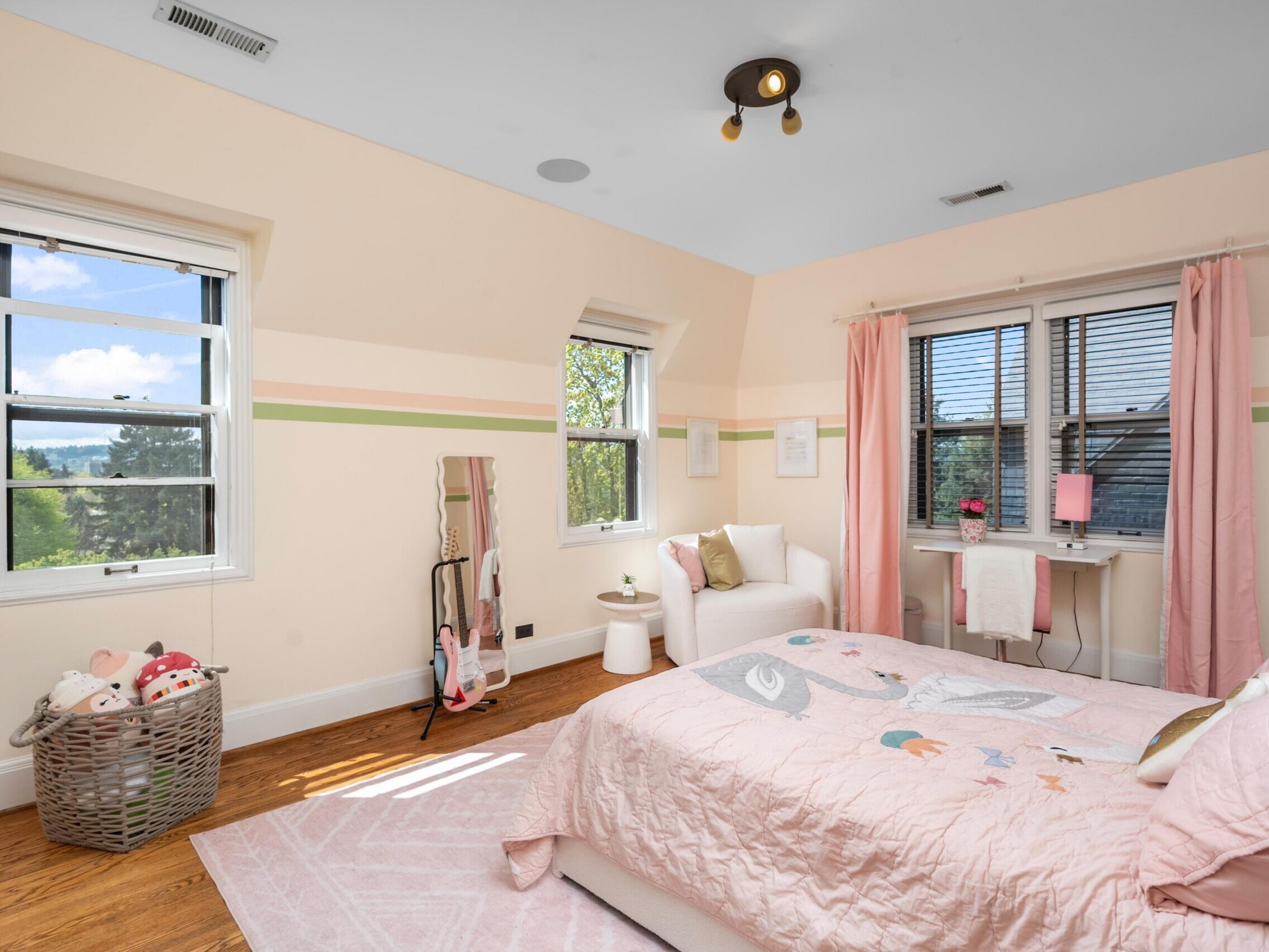 A bright, cozy bedroom with light pink decor, a twin bed with a pink comforter, a white chair, a standing mirror, a laundry basket with plush toys, and large windows letting in natural light.