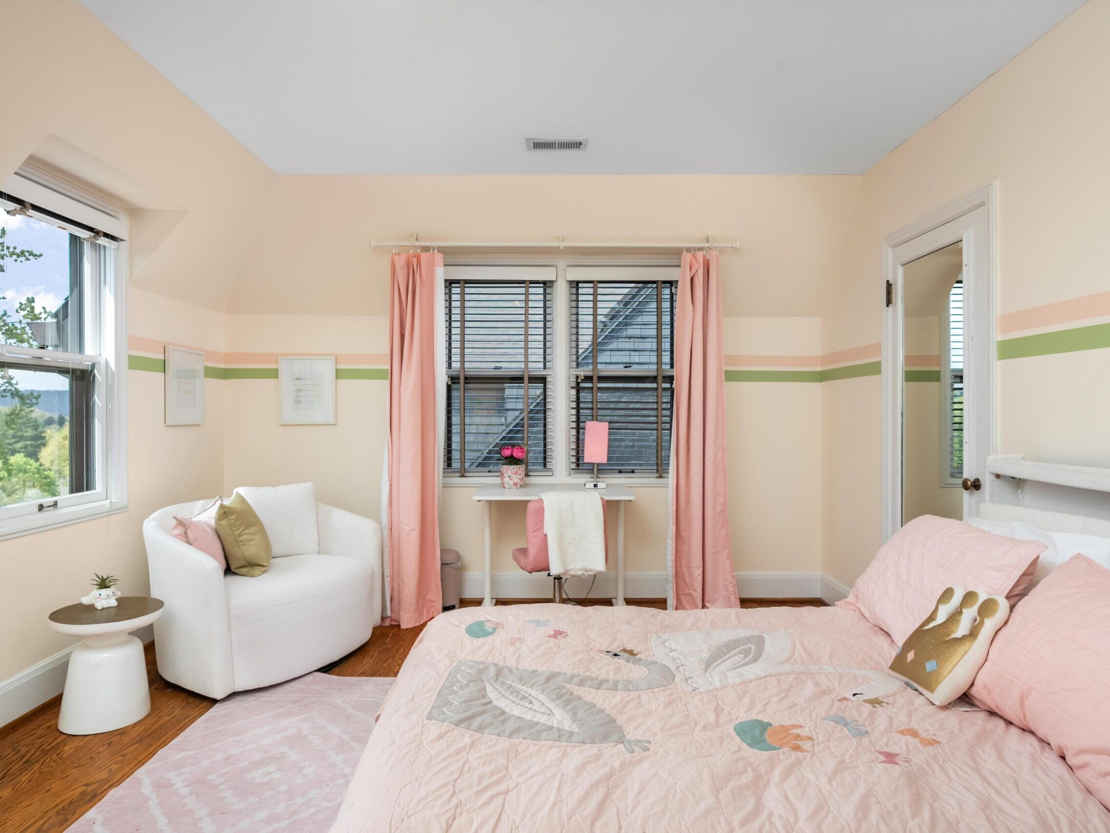 A cozy bedroom with pastel walls and green and pink stripes, featuring a bed with a swan-themed comforter, a small desk by double windows with pink curtains, and a white armchair near another window.