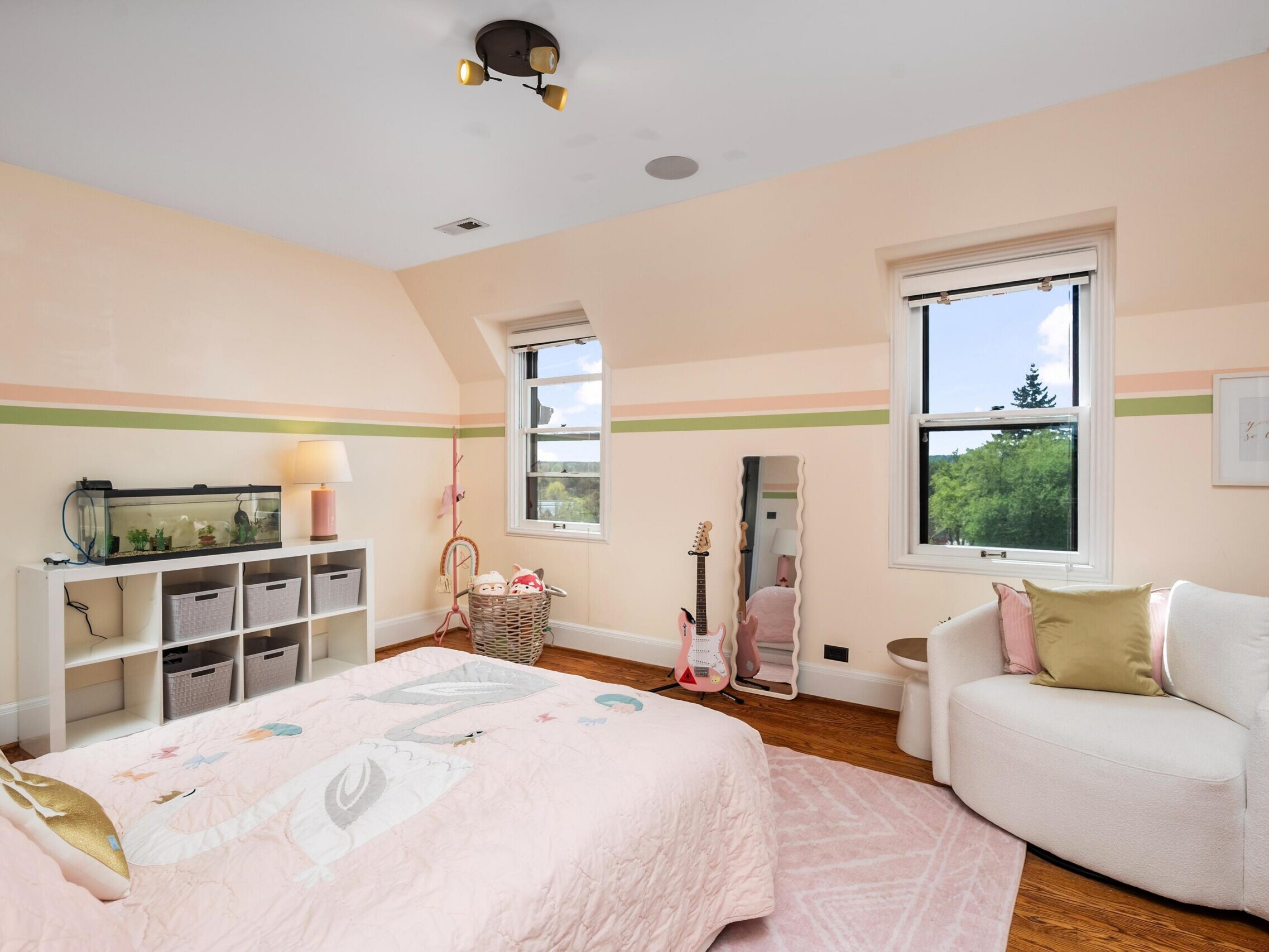 A cozy bedroom with light peach walls, a bed with a pink unicorn-themed blanket, a white armchair, a standing mirror, two windows, and a shelf with bins and a small aquarium. A guitar and basket sit by the wall.