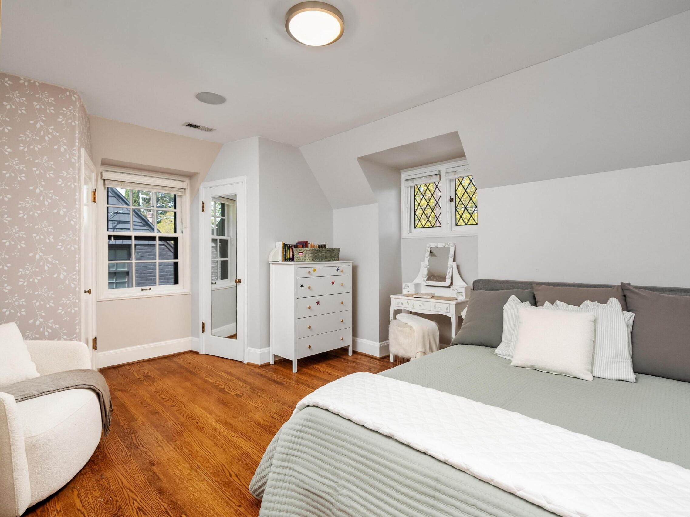 A cozy bedroom with wood floors, a gray bed with white bedding, a white dresser, a vanity table with a mirror, and patterned wallpaper on one wall. Windows let in natural light and a modern armchair sits in the corner.
