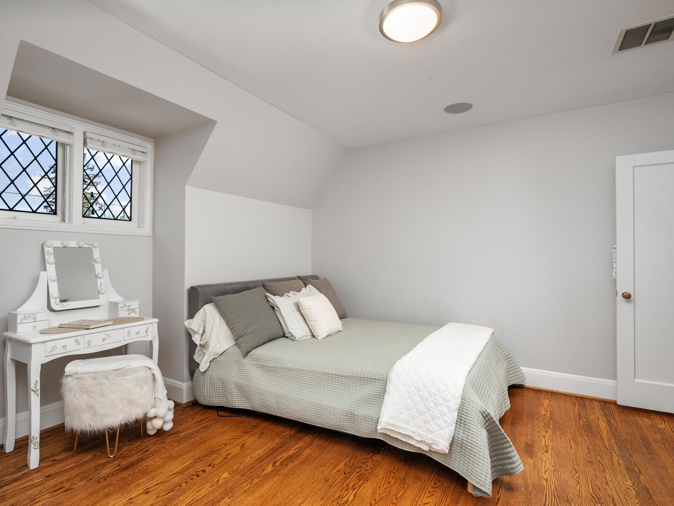 A cozy bedroom with a bed covered in green and white bedding, decorative pillows, and a white vanity table with a mirror and fur stool. The room has hardwood floors, white walls, and a window with diamond-shaped panes.