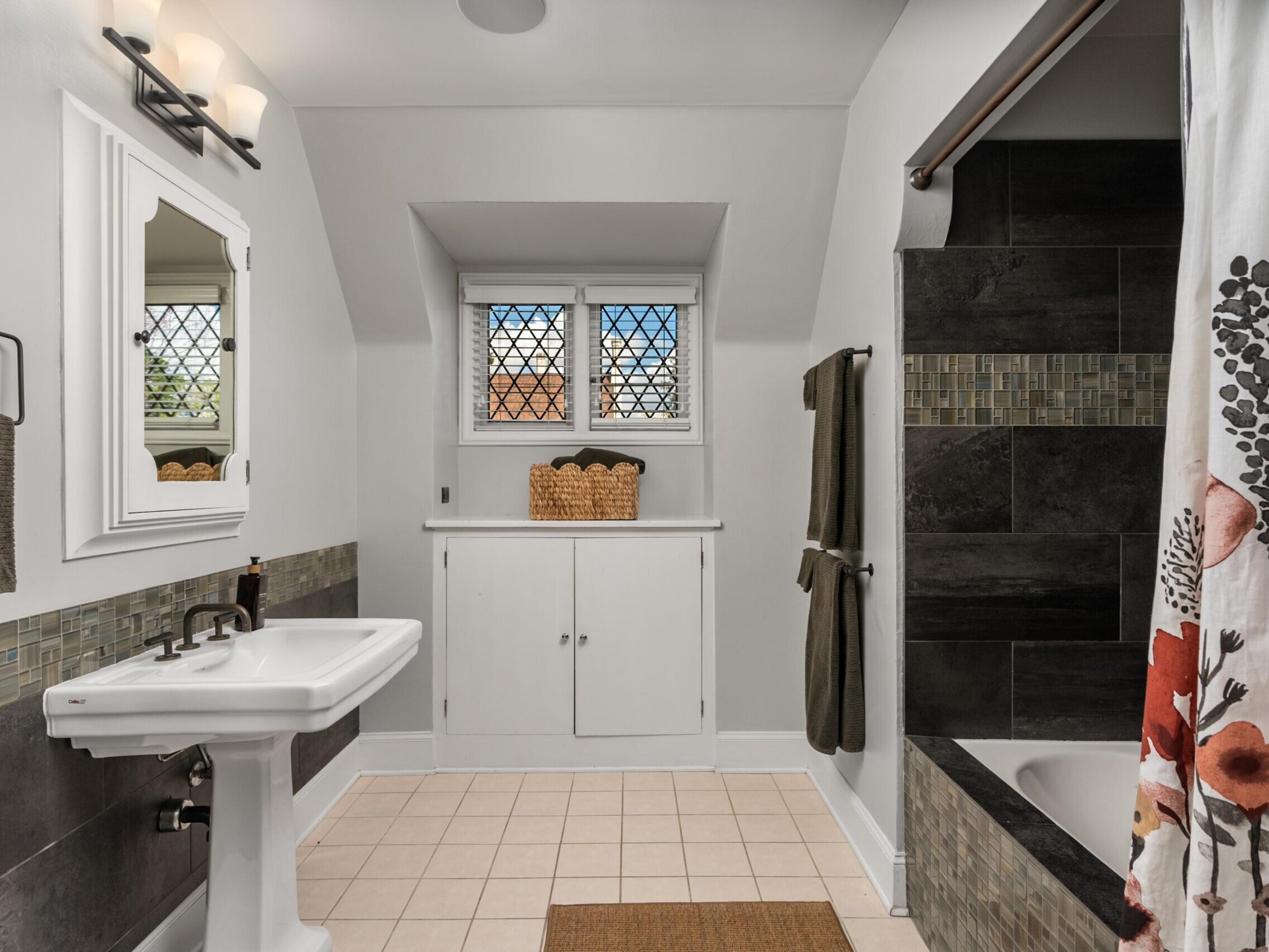 A bright bathroom with a pedestal sink, wall mirror, towel racks, a bathtub with a floral shower curtain, mosaic tile accents, and a window above built-in cabinets with baskets.