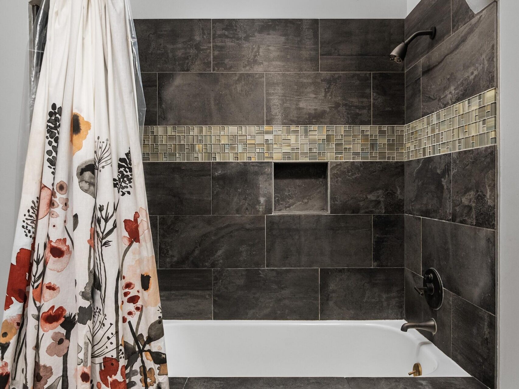 A bathroom with a bathtub surrounded by dark gray tile, a band of mosaic accent tiles, and a shower curtain featuring a floral pattern in red, orange, and gray tones.