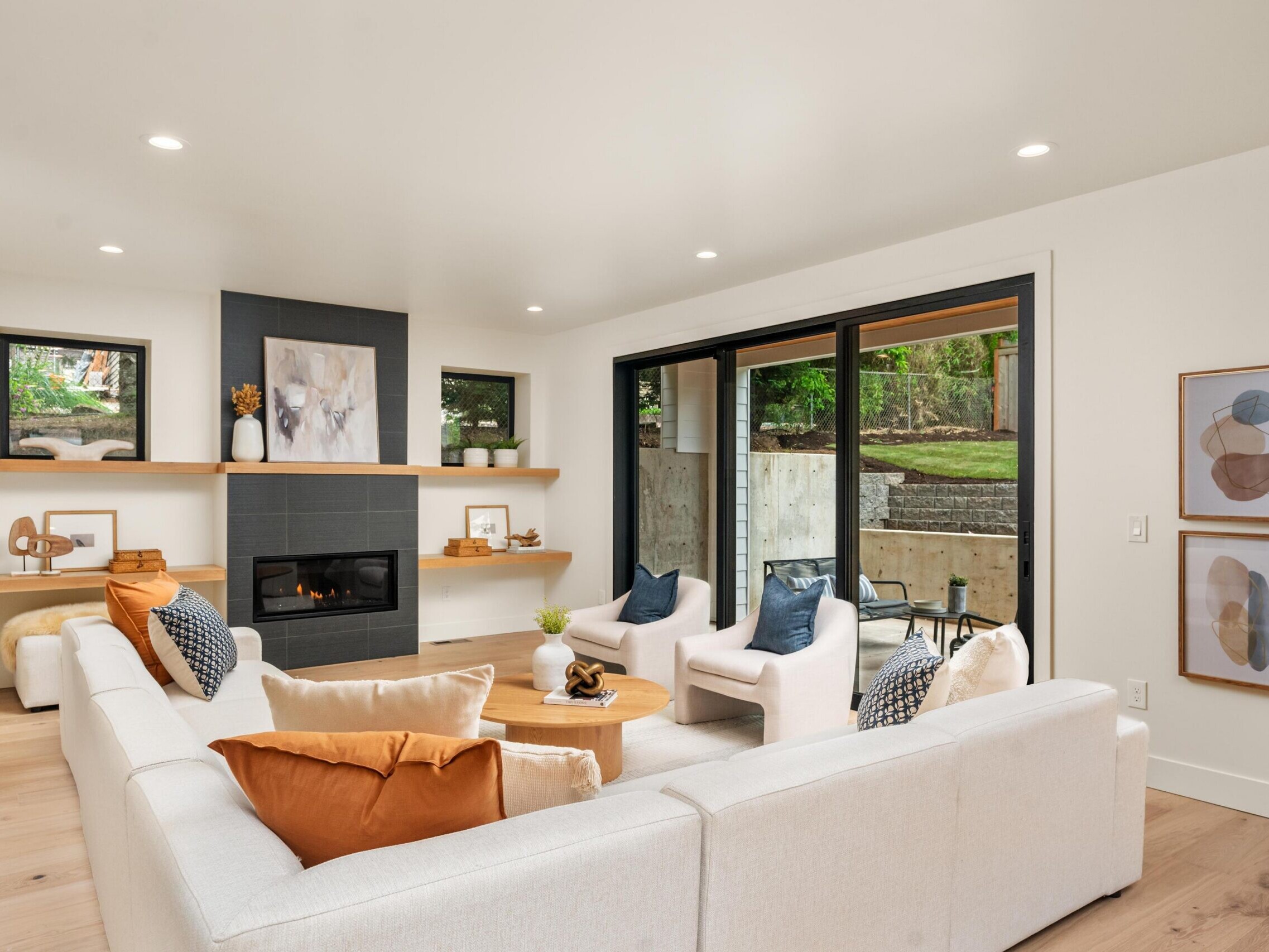 A modern living room with a white sectional sofa, orange and beige pillows, a black fireplace, wooden shelves, abstract art, and large sliding glass doors overlooking a patio and greenery.