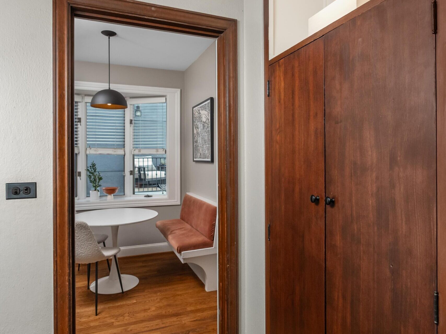 View through a doorway into a dining nook with a round white table, brown leather banquette seating, a black pendant light, and large window. Foreground shows wood flooring and dark wooden built-in cabinets.