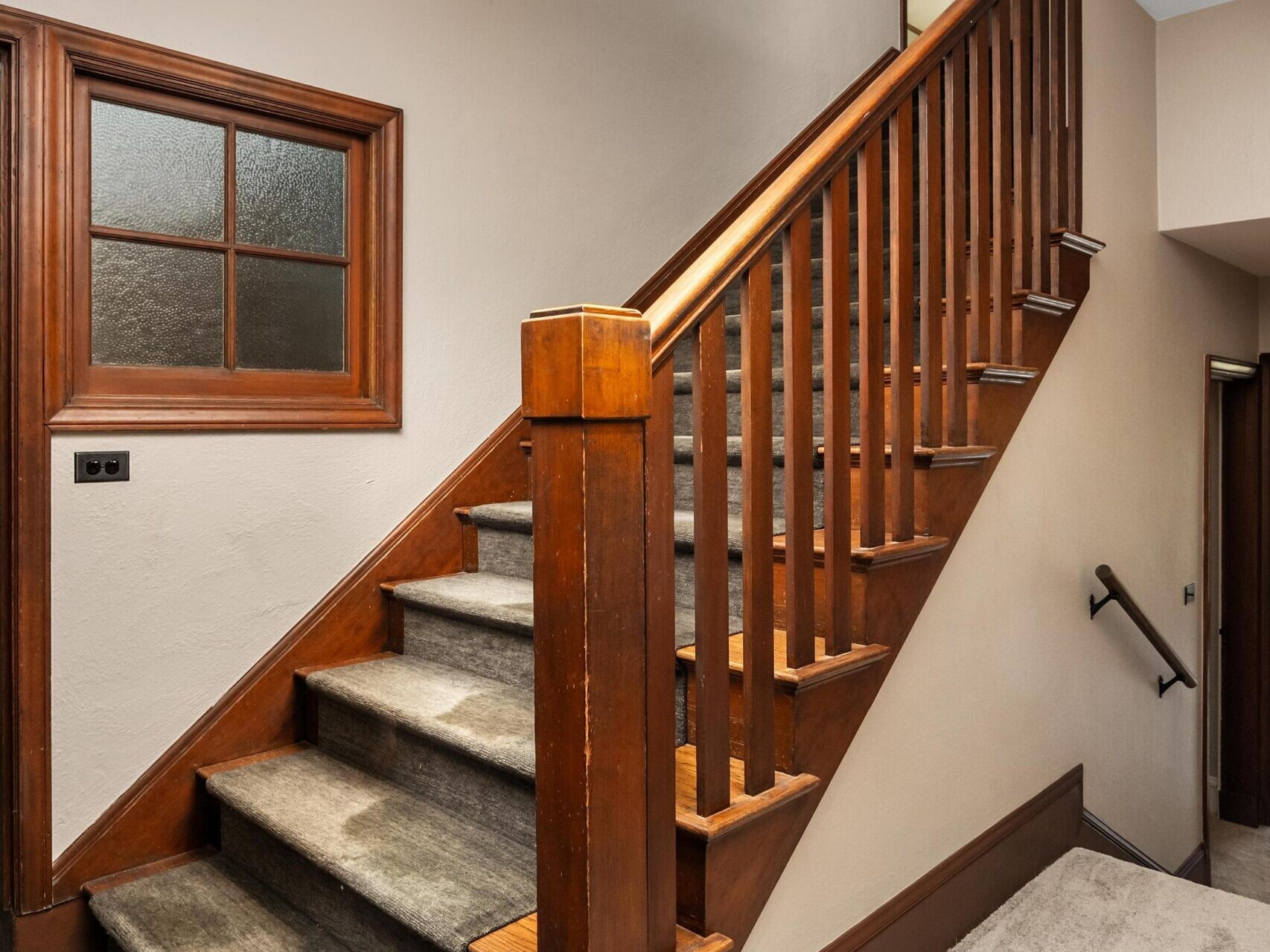 A wooden staircase with carpeted steps, beige walls, and a wooden handrail, located in a residential hallway with light carpet flooring and a small window on the left wall.