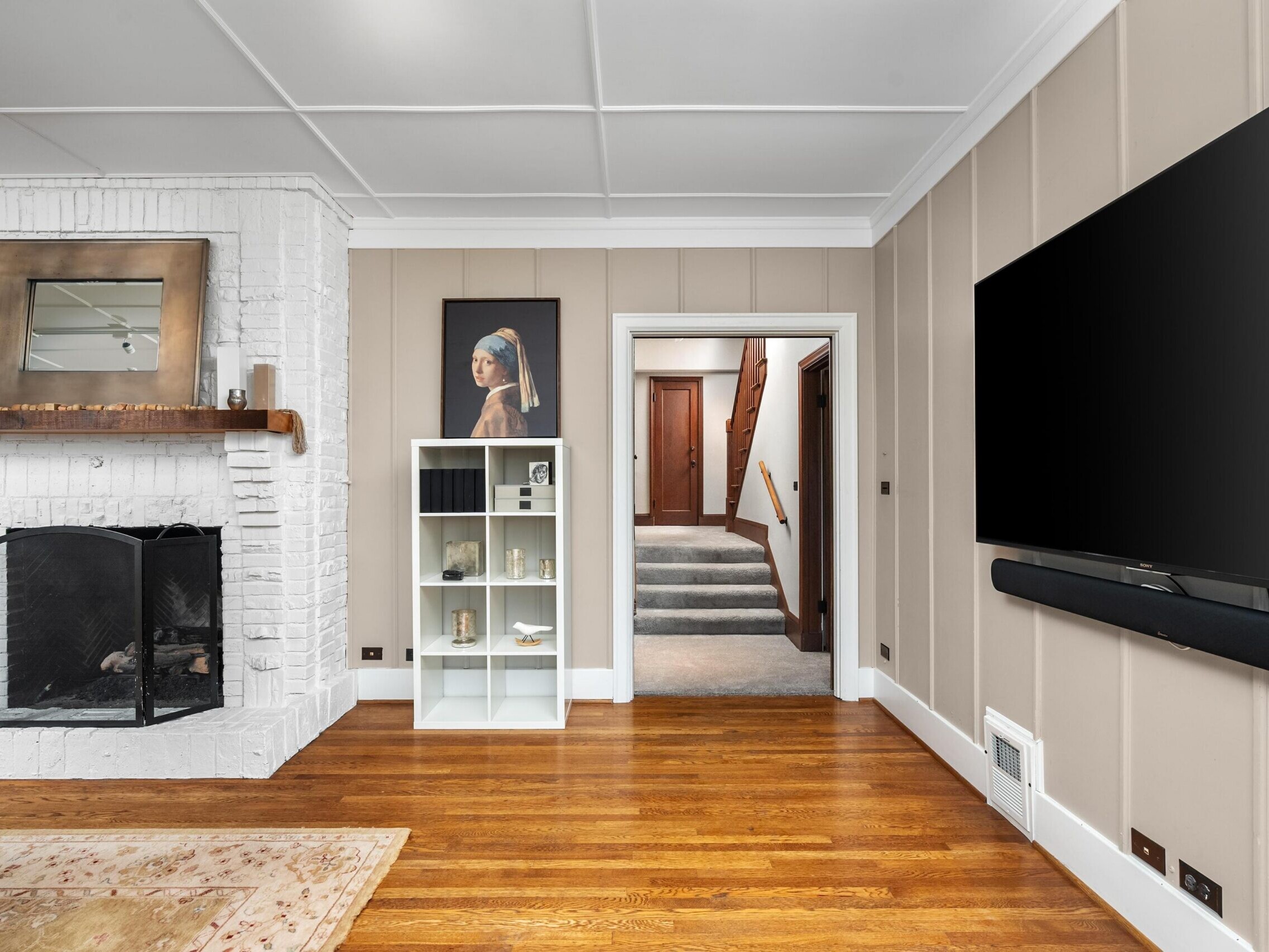 A modern living room with hardwood floors, a large wall-mounted TV, a white brick fireplace with a wood mantel, a bookshelf with decor, and a hallway leading to stairs.