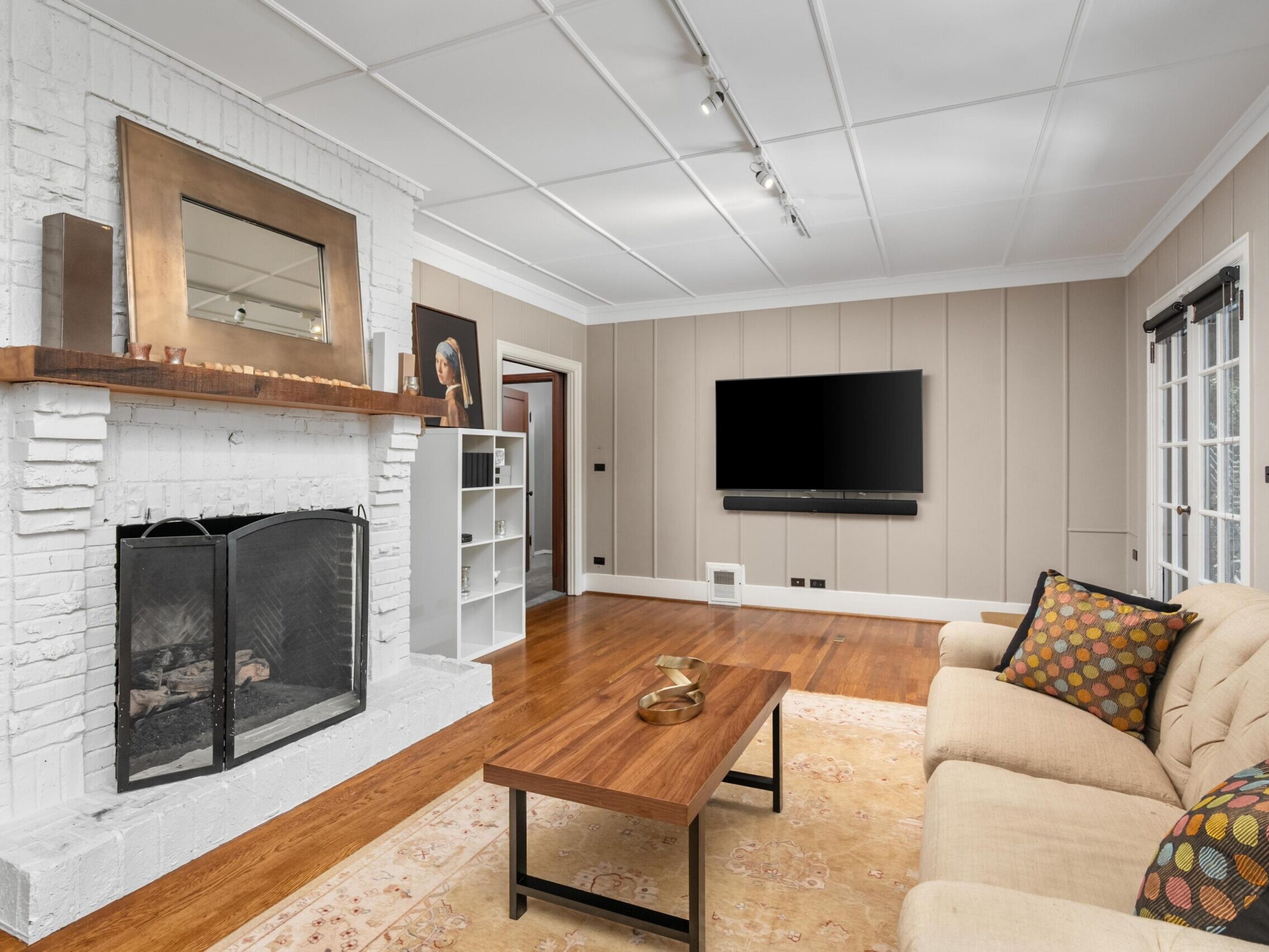 A cozy living room with a beige couch, patterned pillows, wooden coffee table, white brick fireplace with a mirror above it, wall-mounted TV, and hardwood floors. A shelf stands near the corner, and windows let in natural light.