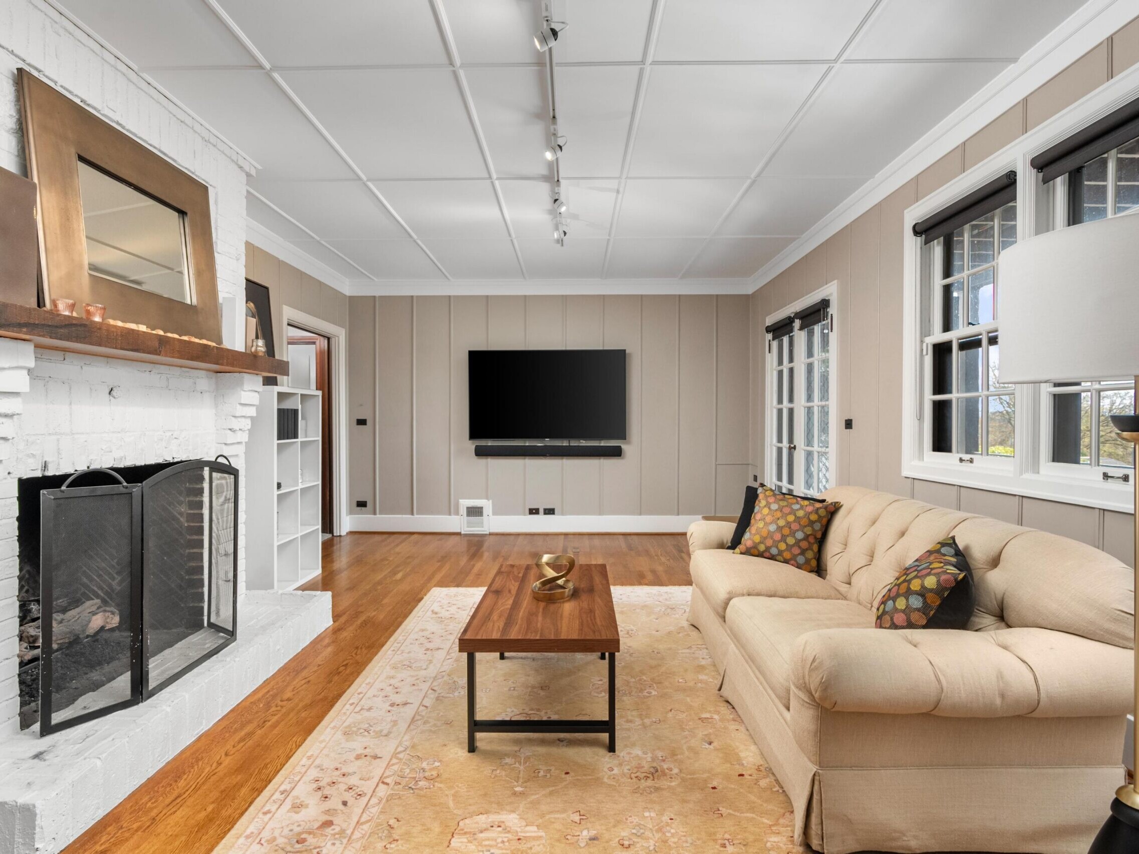 A cozy living room with a beige sofa, a wooden coffee table, a white brick fireplace with a mirror above, a large wall-mounted TV, and windows letting in natural light. The room features wood floors and neutral walls.
