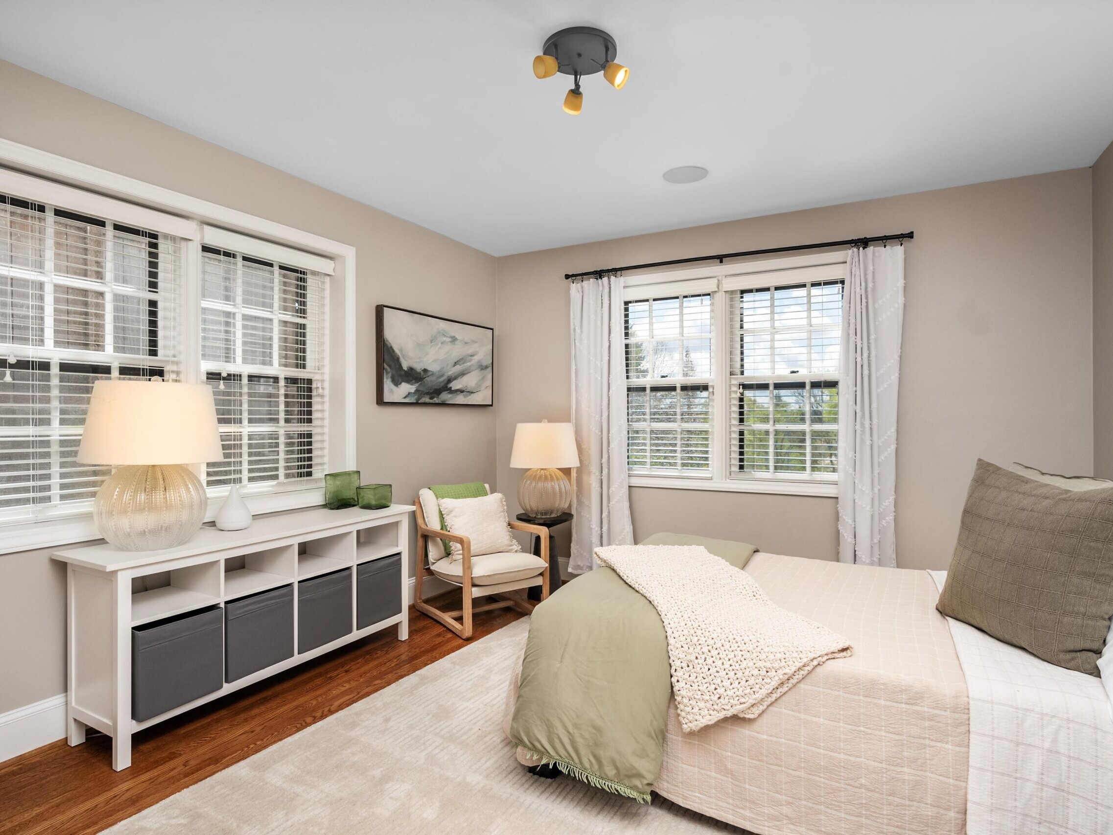A cozy bedroom with two large windows, a neatly made bed with green and beige bedding, a white console table with lamps, an armchair with a pillow, wall art, and wooden floors. Natural light brightens the room.