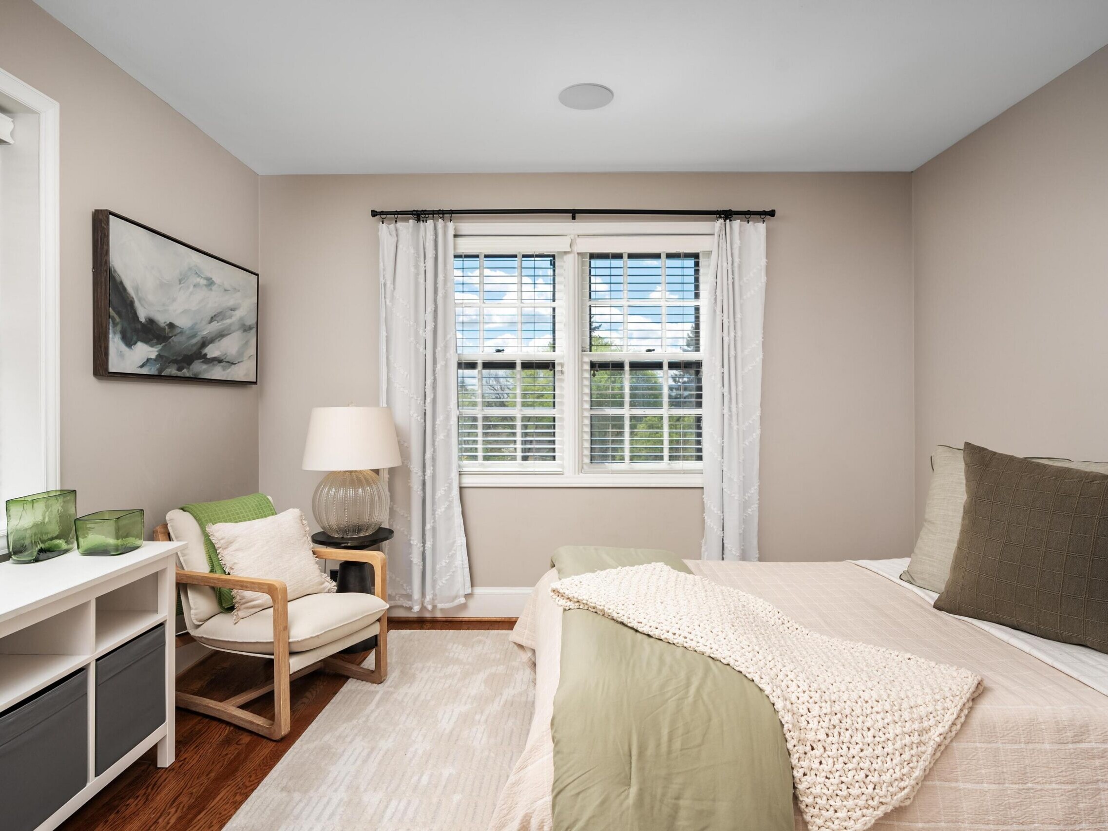 A cozy bedroom with a bed, green and beige bedding, a white rug, a wooden chair with a green pillow, a lamp, and a window with white curtains letting in natural light. Decor includes abstract wall art and glass vases.