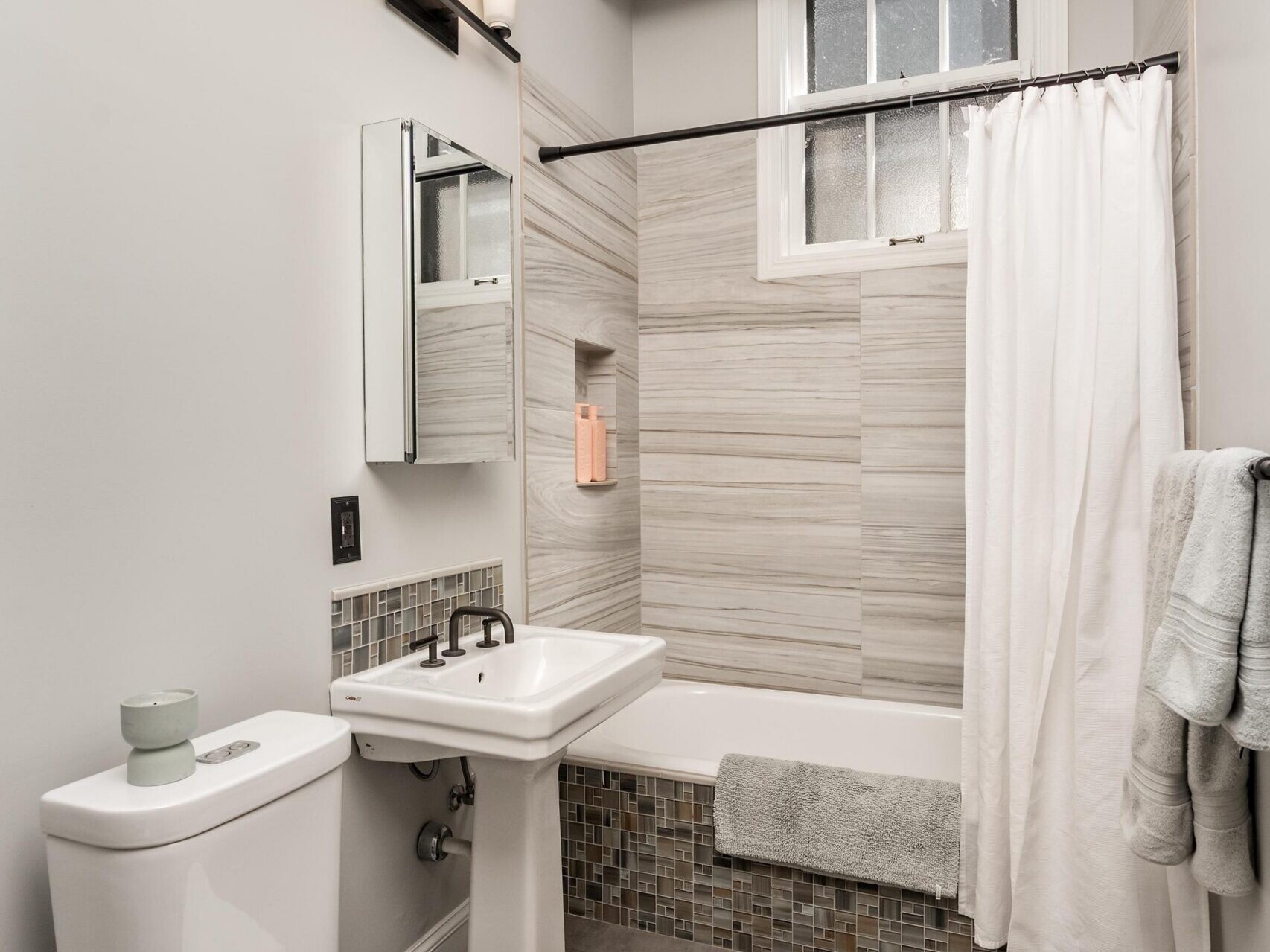 A modern bathroom with a white toilet, pedestal sink, and a bathtub with a light tile surround and white shower curtain. Above the sink is a mirror and three-light fixture. Towels hang on the wall; the floor is tiled gray.