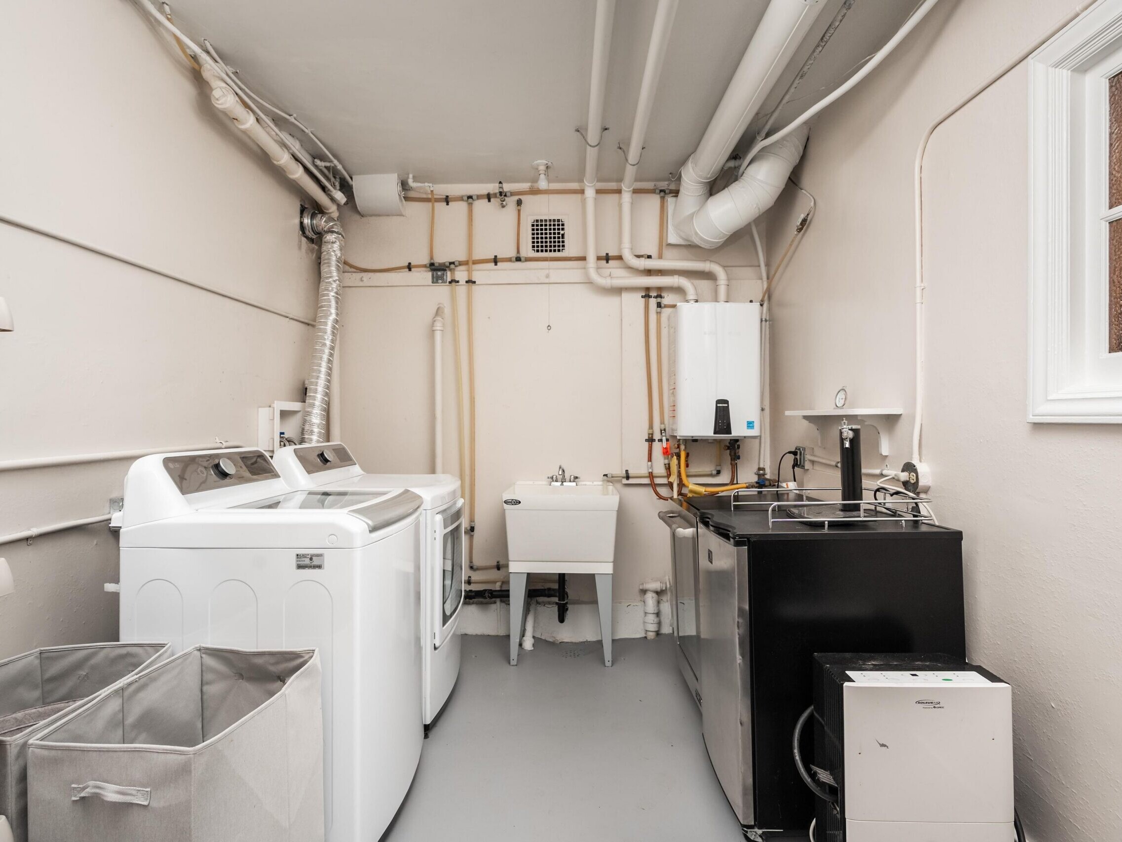 A clean laundry room with a white washer and dryer, utility sink, metal shelving, storage bins, and exposed pipes on the walls. There is a window on the right and a black utility cabinet with a faucet.