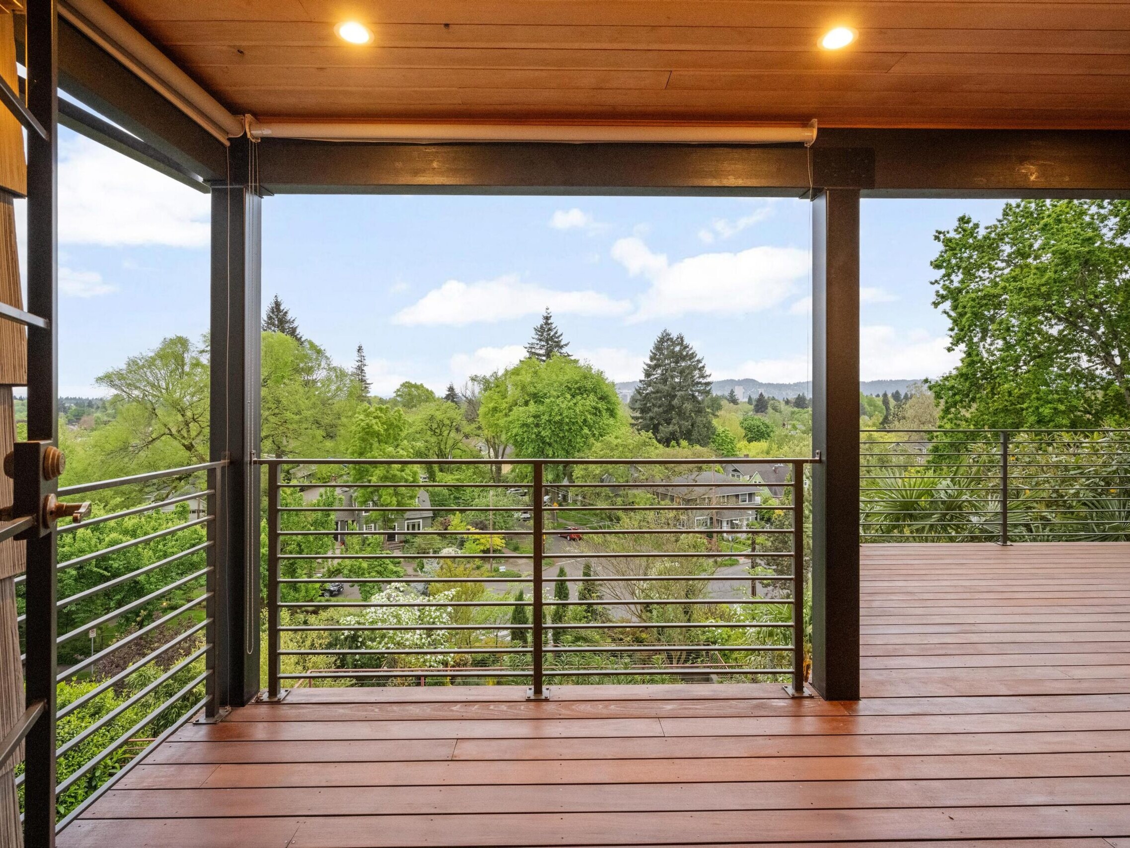 A spacious wooden balcony with metal railings overlooks a lush green landscape with trees and distant hills under a partly cloudy sky. Warm ceiling lights illuminate the covered area.
