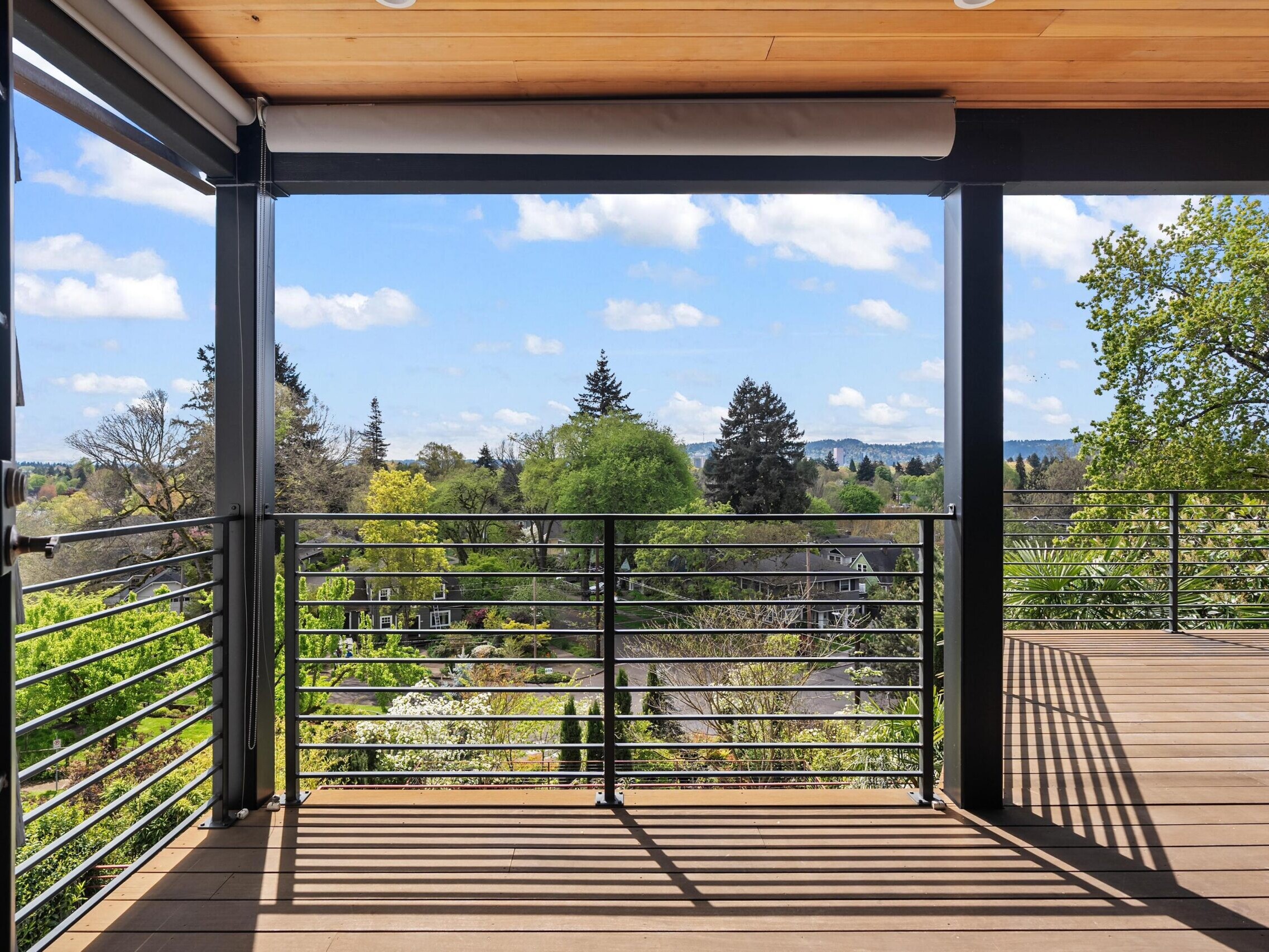 A spacious balcony with metal railings offers a scenic view of green trees, rooftops, and distant hills under a blue sky with scattered clouds. The deck has a wooden floor and ceiling.