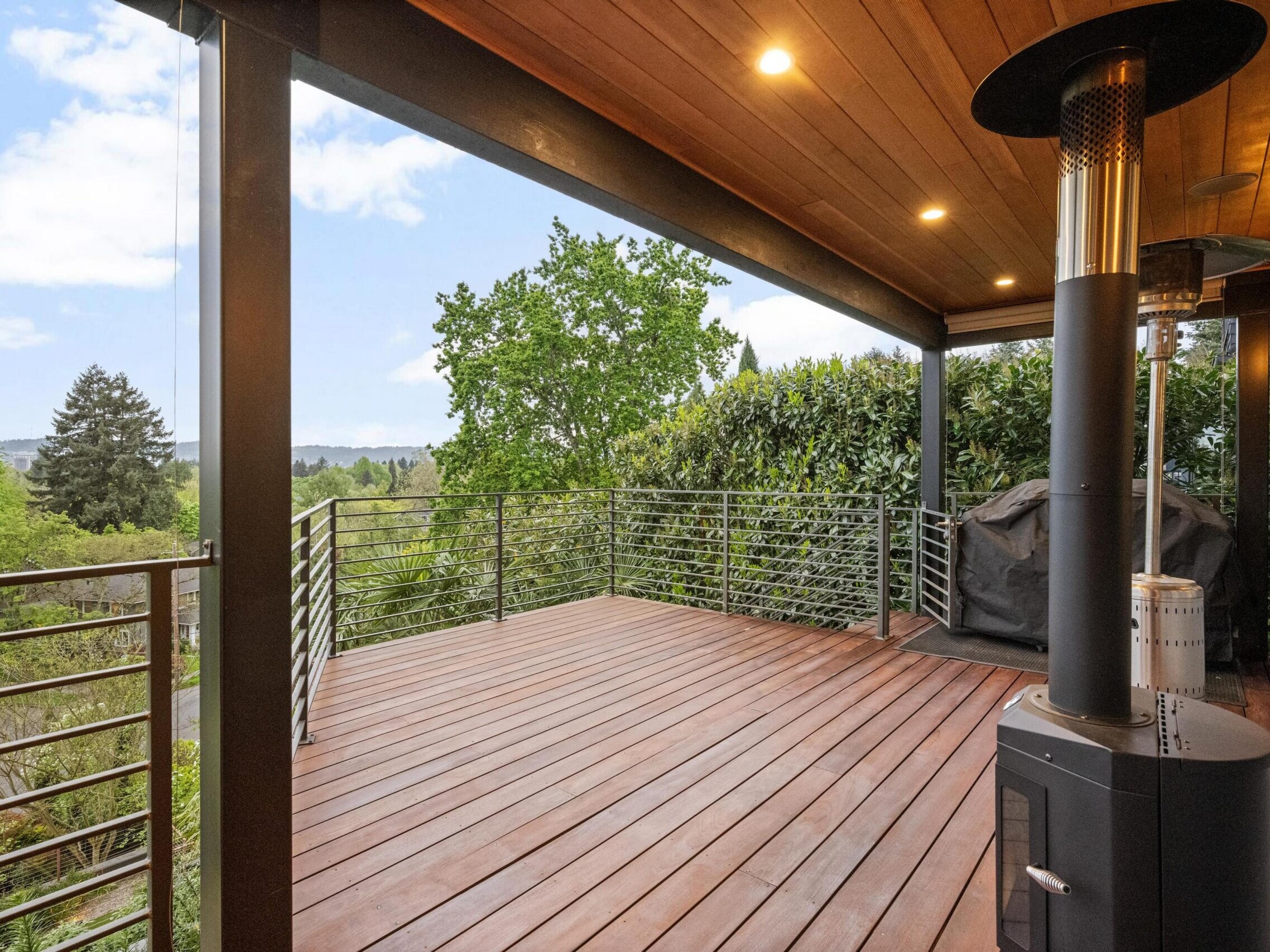A modern wooden deck with metal railings, ceiling lights, and an outdoor heater overlooks lush green trees and a clear sky. A covered grill sits in one corner of the spacious patio.