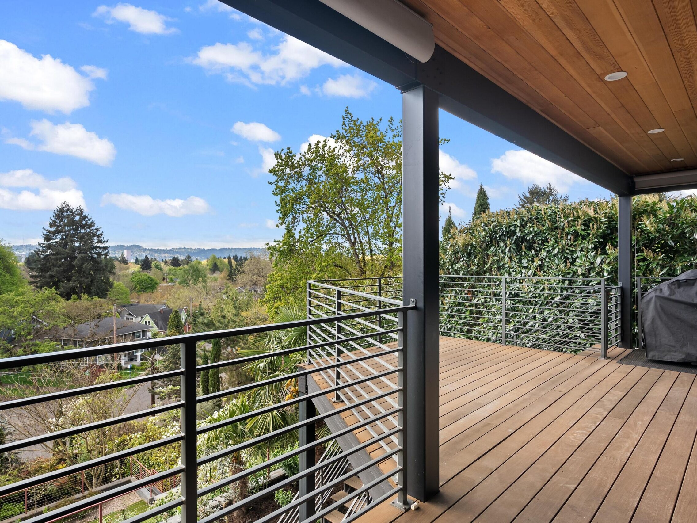A spacious wooden deck with metal railings overlooks a lush, green neighborhood with trees and houses below. The sky is blue with scattered clouds, creating a bright and inviting atmosphere.